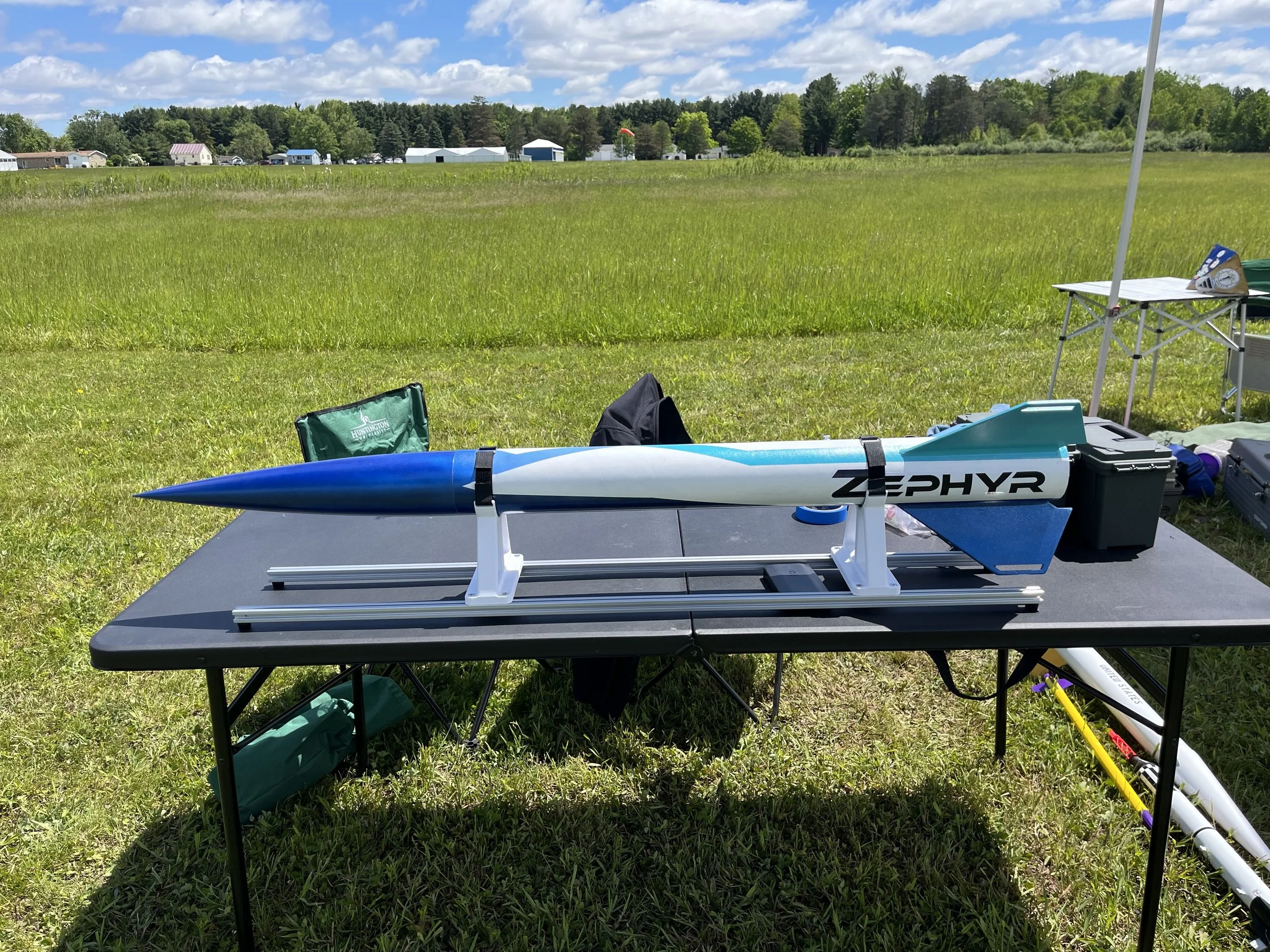 Model rocket on a launch pad set up on a table outdoors in a grassy field with cloudy sky, with trees and some buildings in the background.