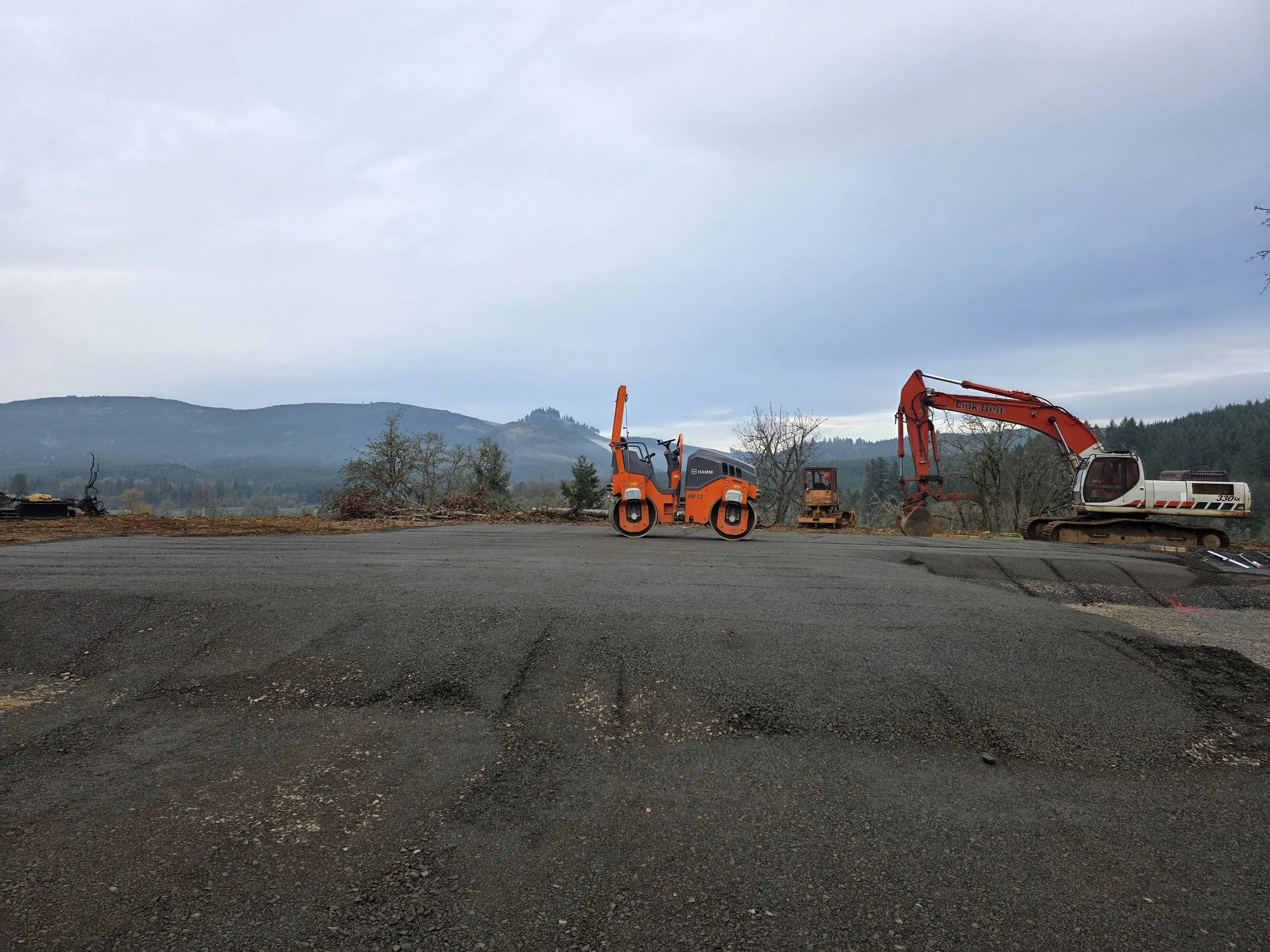 Construction site with heavy equipment and machinery in a rural area with mountains in the background and a cloudy sky.