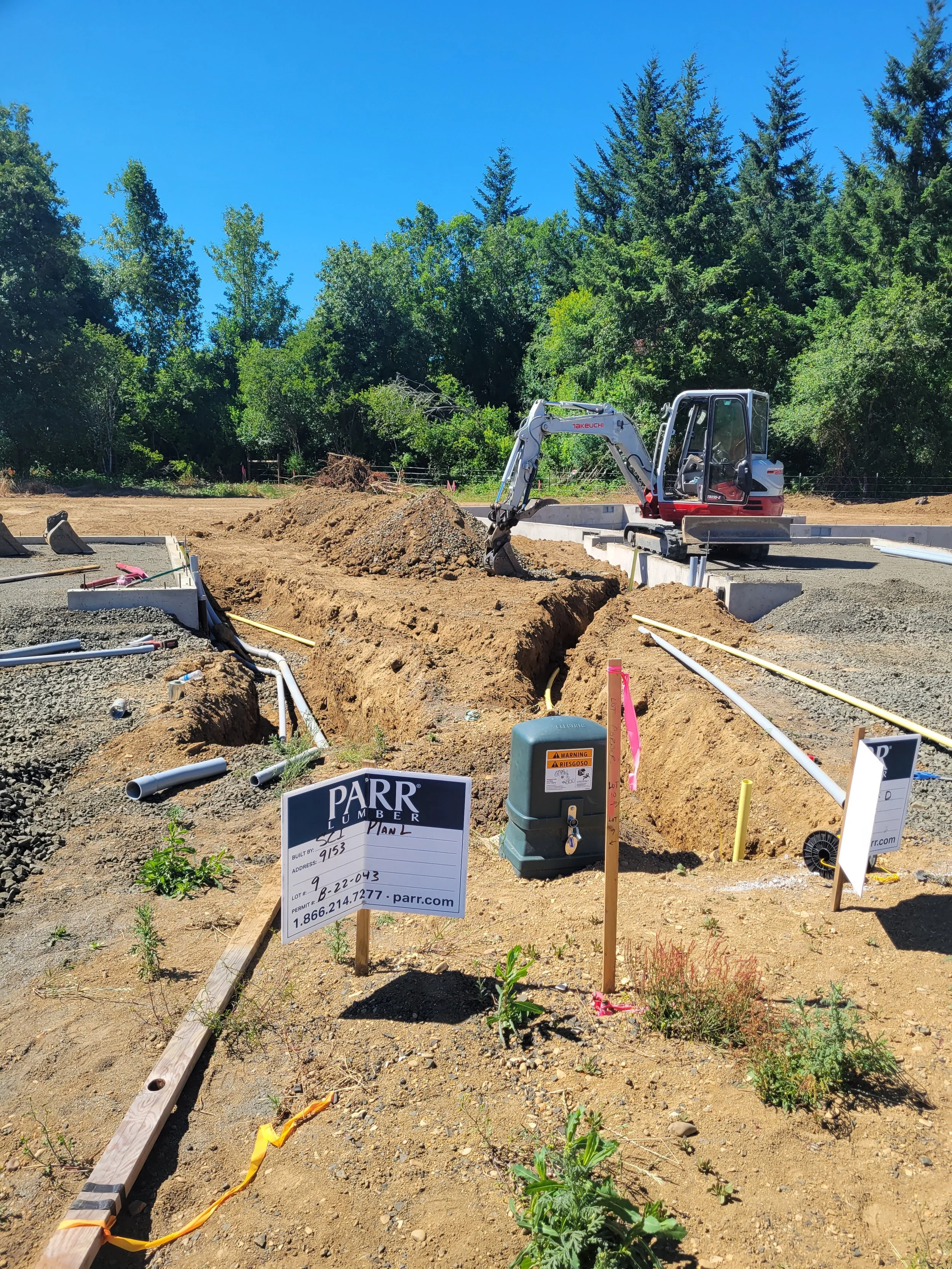Construction site with a small excavator working on a dirt area, surrounded by trees and a clear blue sky, with construction signs and pipes on the ground.