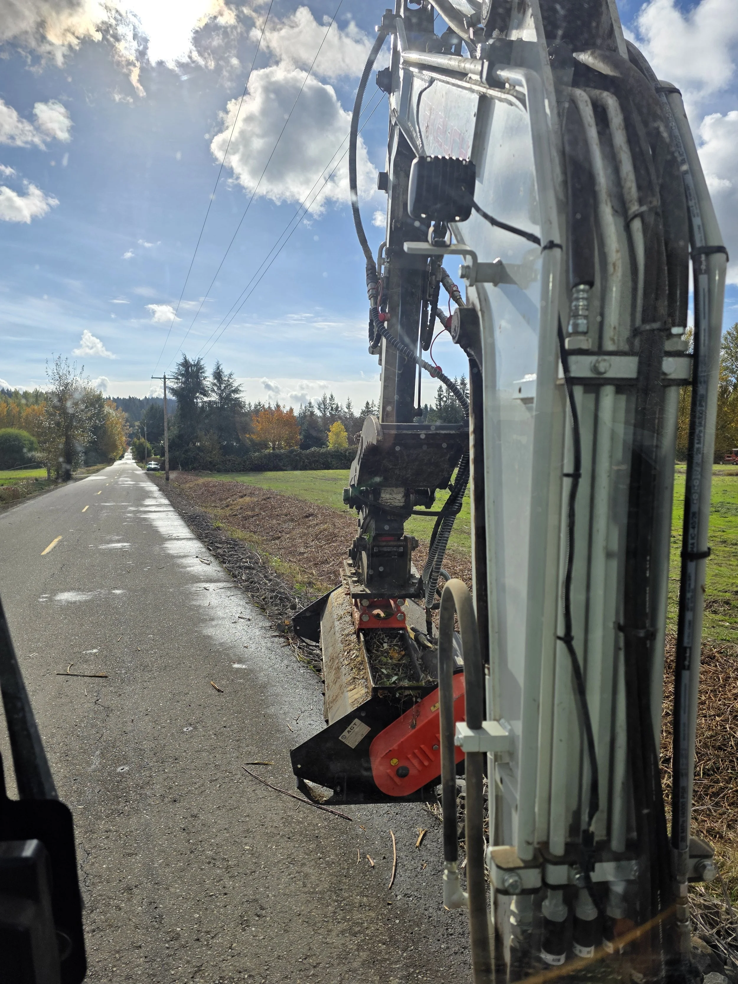 Close-up view from inside a construction vehicle showing machinery and hydraulic components on the right side, with a road, trees with fall foliage, and cloudy sky in the background. Salem Oregon