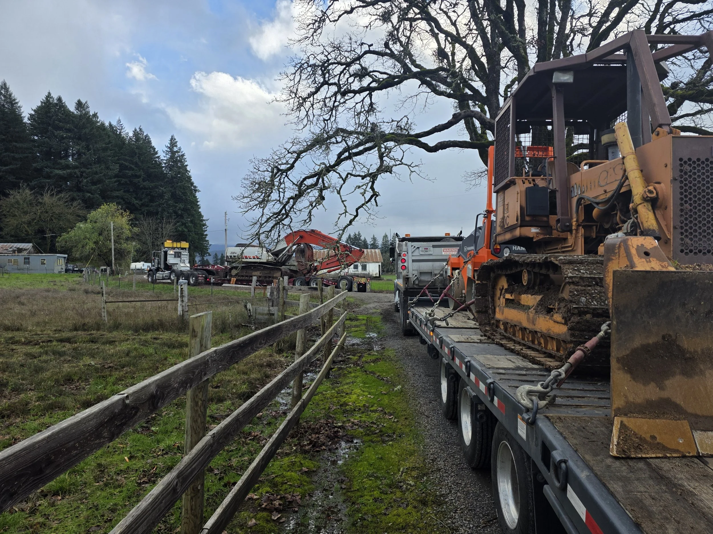 Large construction vehicle on a flatbed trailer, parked on a muddy road with grass and a wooden fence, with a leafless tree and a cloudy sky in the background.