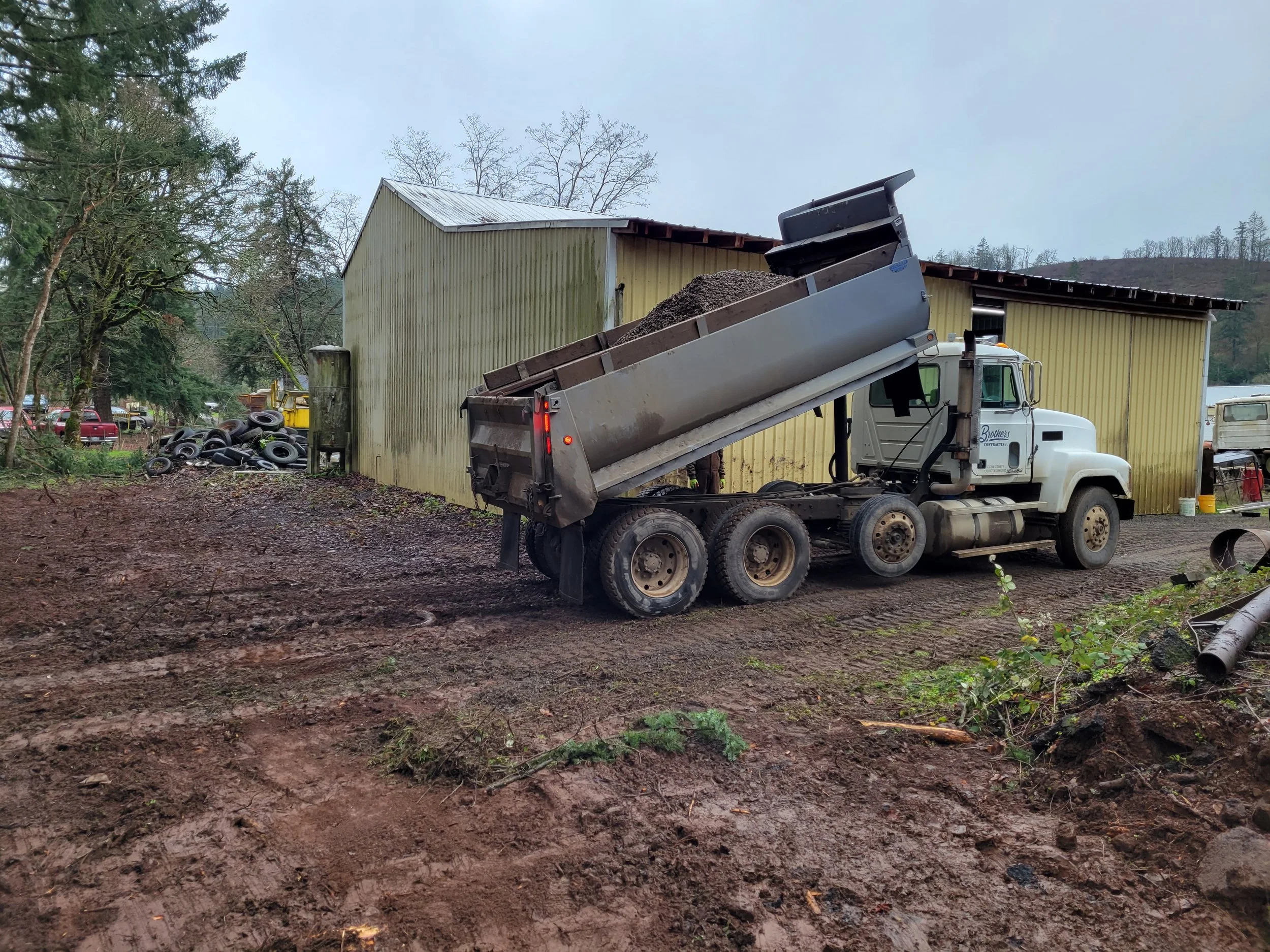 A dump truck with a raised bed dumping gravel or dirt next to a yellow metal building, with trees and hills in the background on a cloudy day.