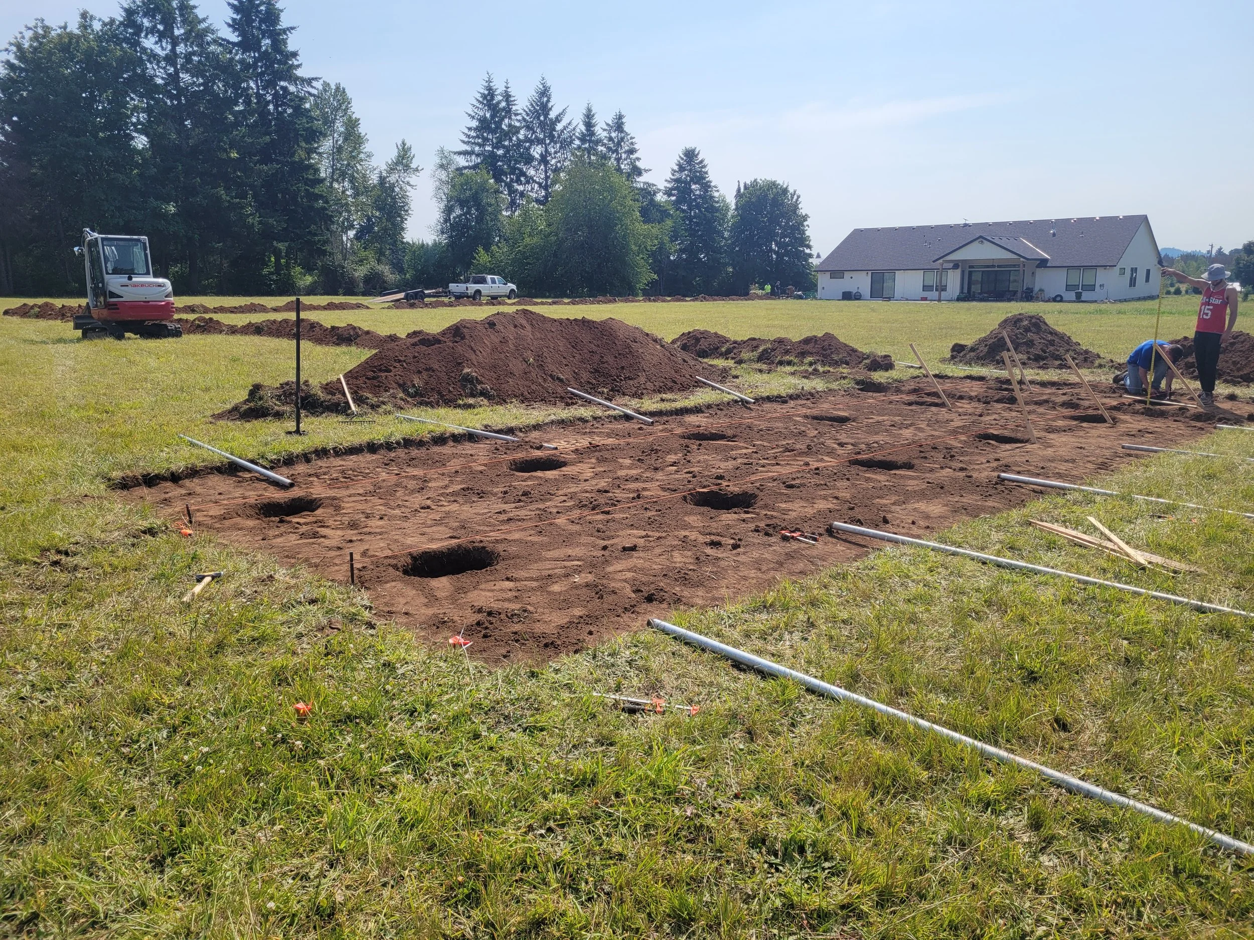 Construction site with dug holes, piles of dirt, and construction workers working on foundation for a new building. There is a small excavator, a white pickup truck, and a modern house in the background under a clear sky.