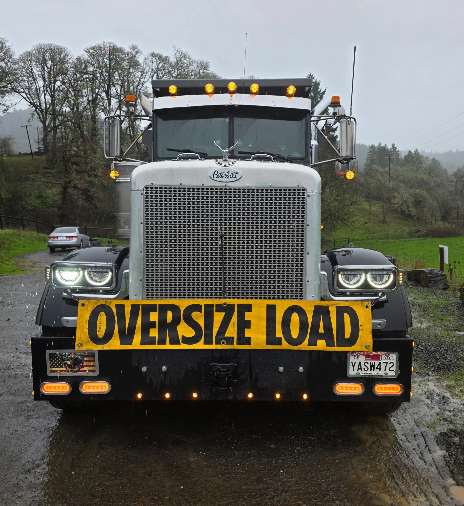 Front view of a large Peterbilt semi-truck with an oversized load sign on the bumper, illuminated headlights, turn signals, and orange warning lights on top, parked outdoors with trees and a cloudy sky in the background. Aumsville Oregon.