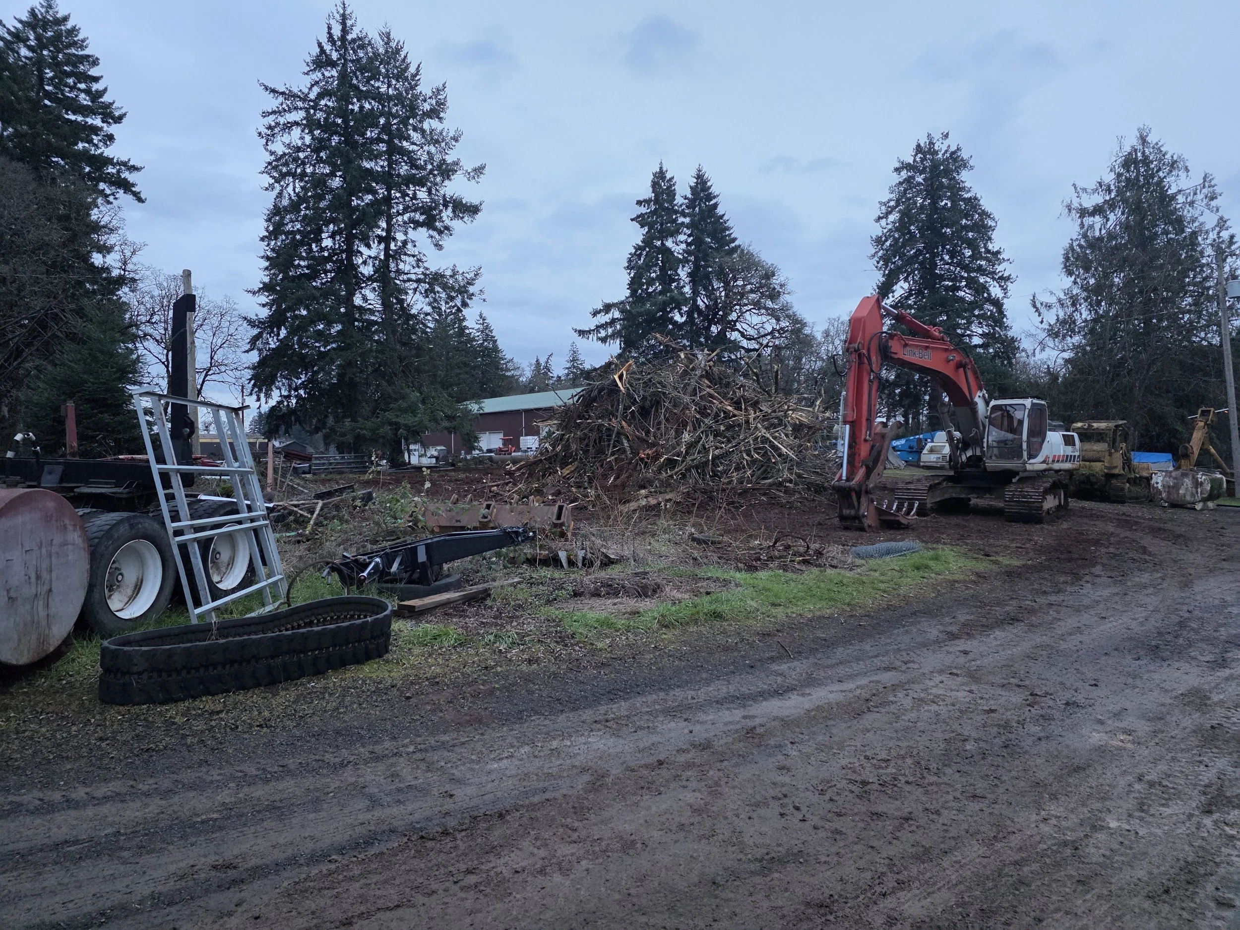 Scene of an outdoor yard with a small excavator, a large pile of wood debris, trees, a barn, and some machinery or trailers, captured during evening or early morning.