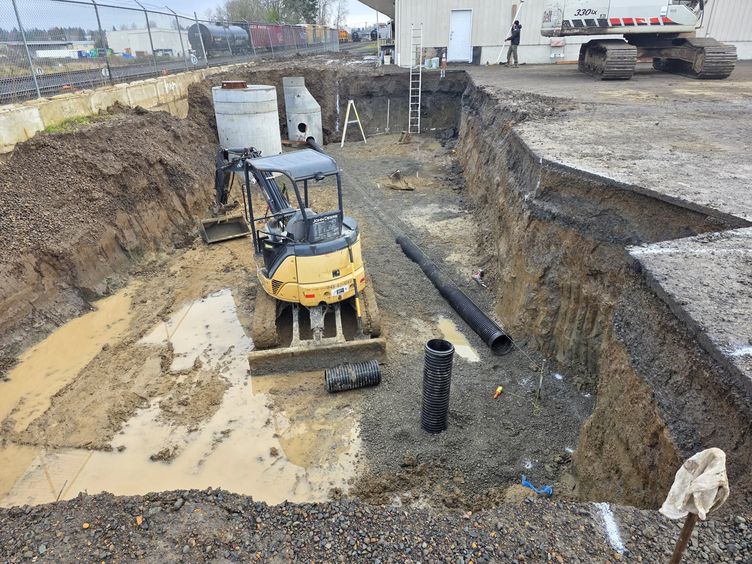 Construction site with a deep excavated trench, a mini excavator, black drainage pipes, and a worker operating a shovel near a building with train tracks in the background. Woodburn Oregon