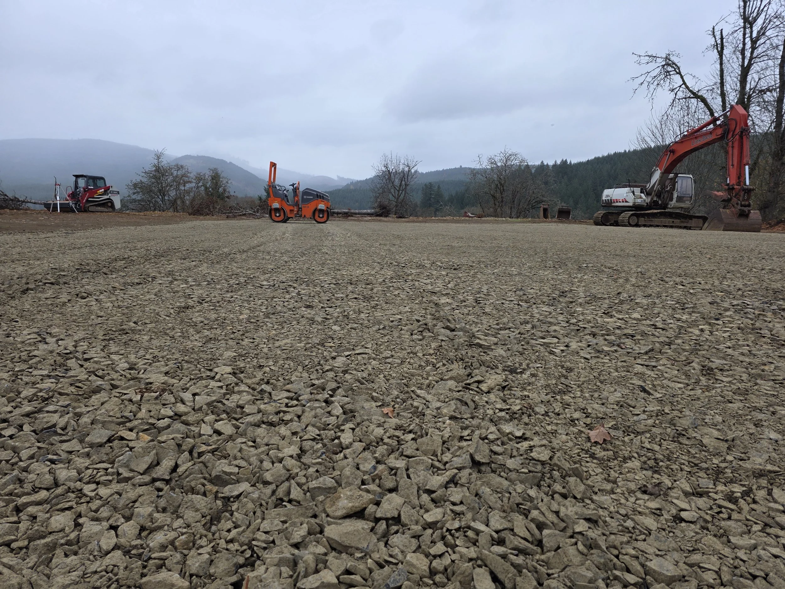A construction site with gravel ground, a small red roller compactor, a large orange backhoe, and a smaller gray machine, with trees, mountains, and cloudy sky in the background.