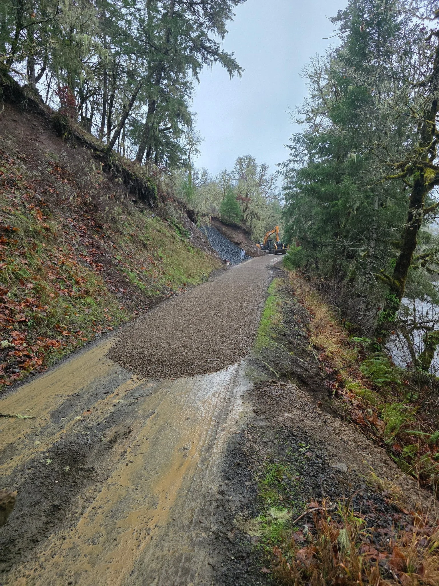 A dirt and gravel trail along a hillside, with construction equipment and workers near a slope, surrounded by trees and overcast sky.