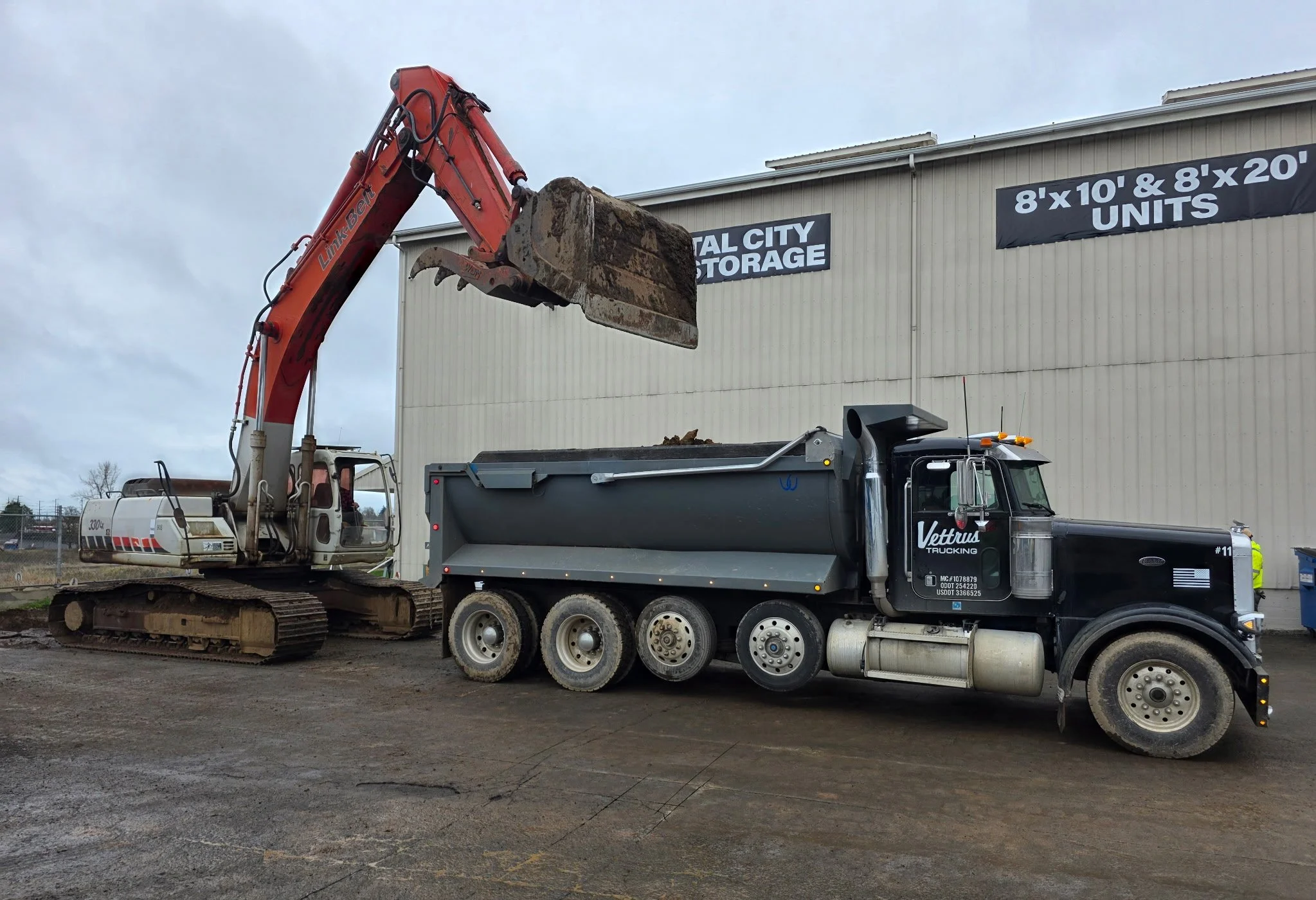 A construction excavator with the arm raised and bucket filled with dirt beside a black dump truck at a storage facility.