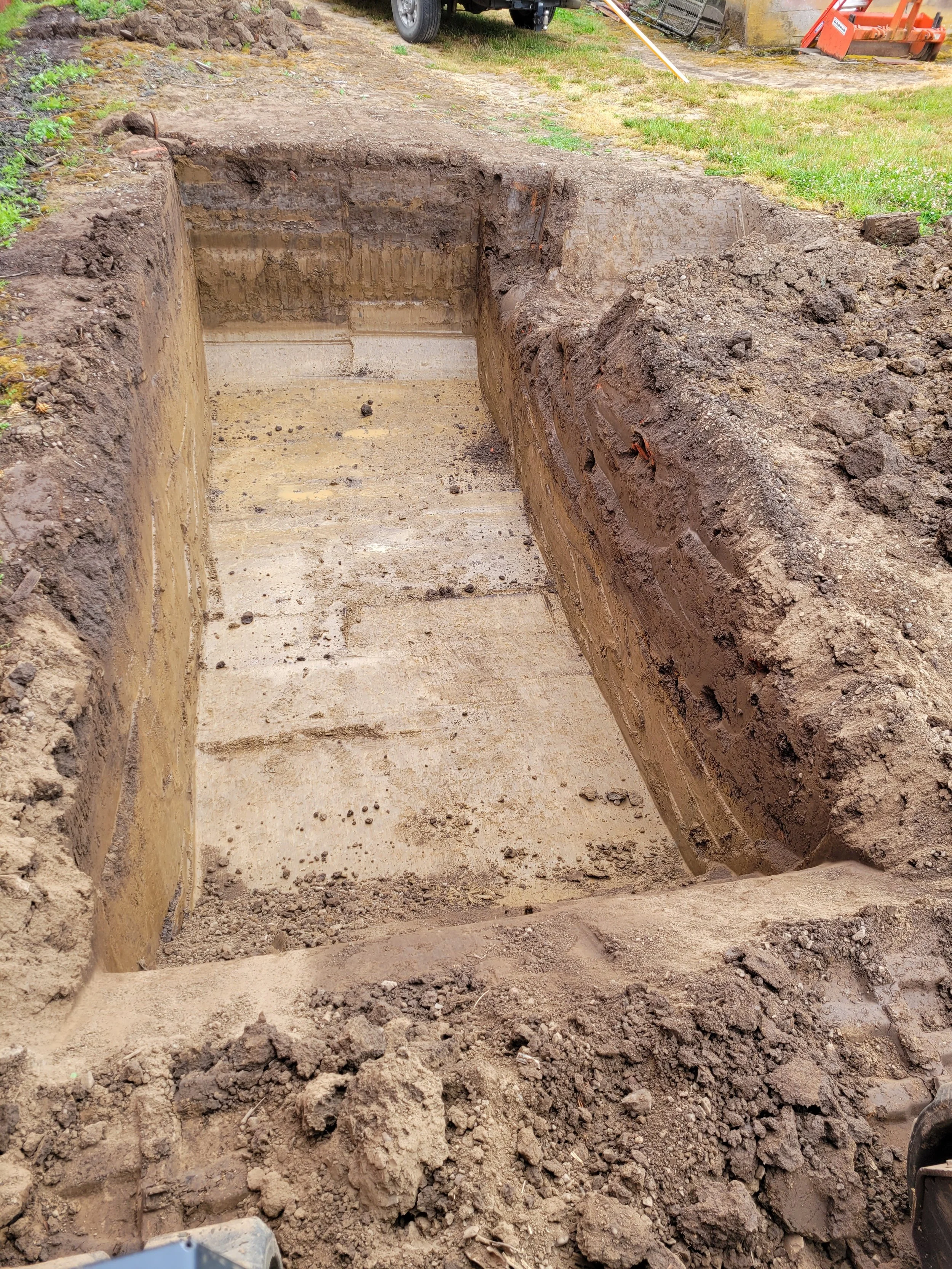 A deep excavated trench at a construction site with exposed soil layers and earth piled around the edges, with some construction equipment and grass in the background.