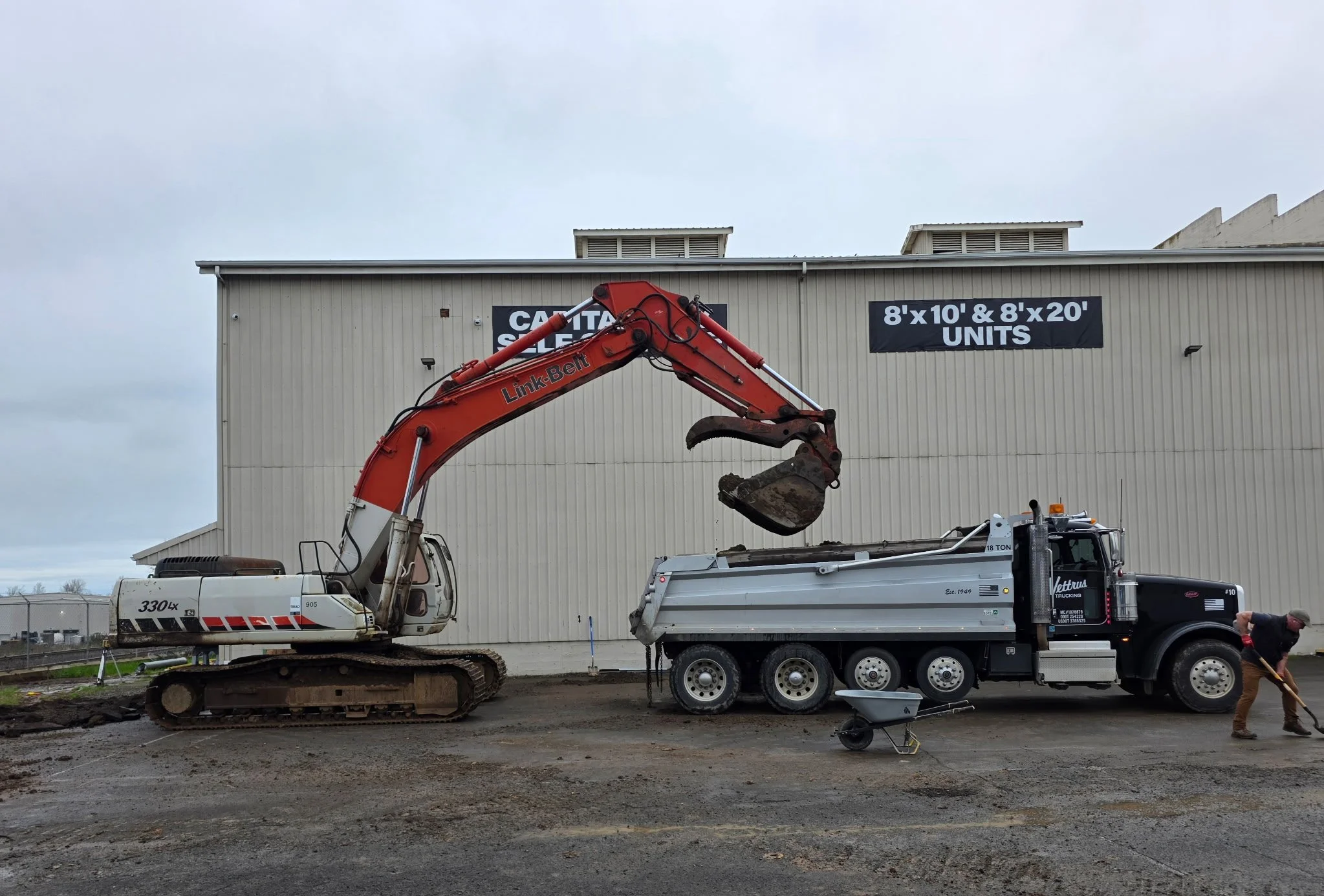 Construction site with excavator and flatbed truck in front of a large industrial building with signage for unit sizes