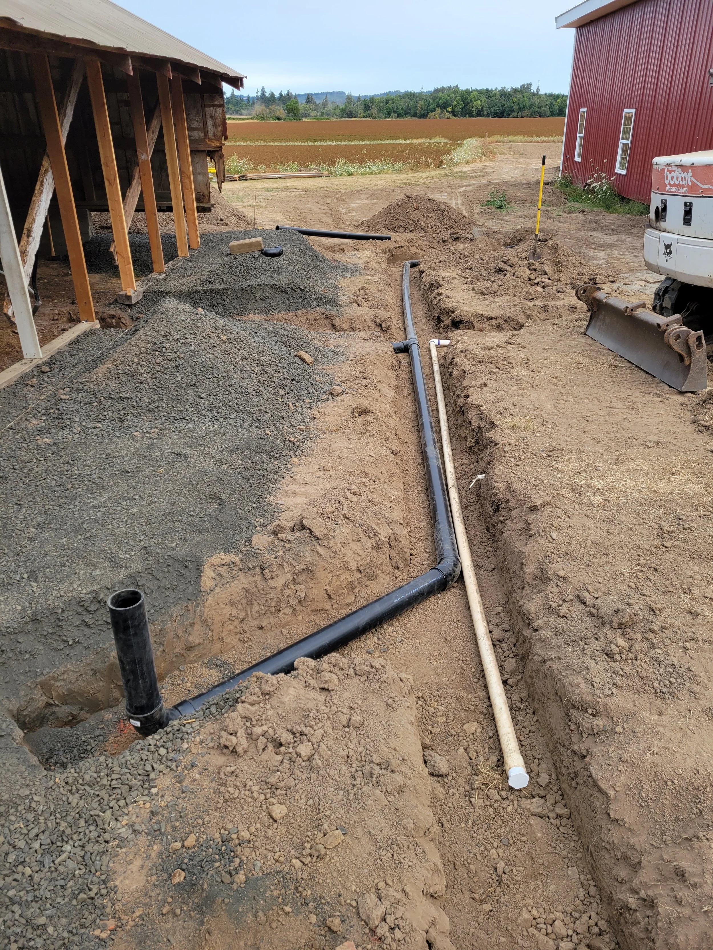 Construction site showing pipes in a trench next to a building, with a pile of dirt and construction tools, in a rural area with fields and trees in the background.