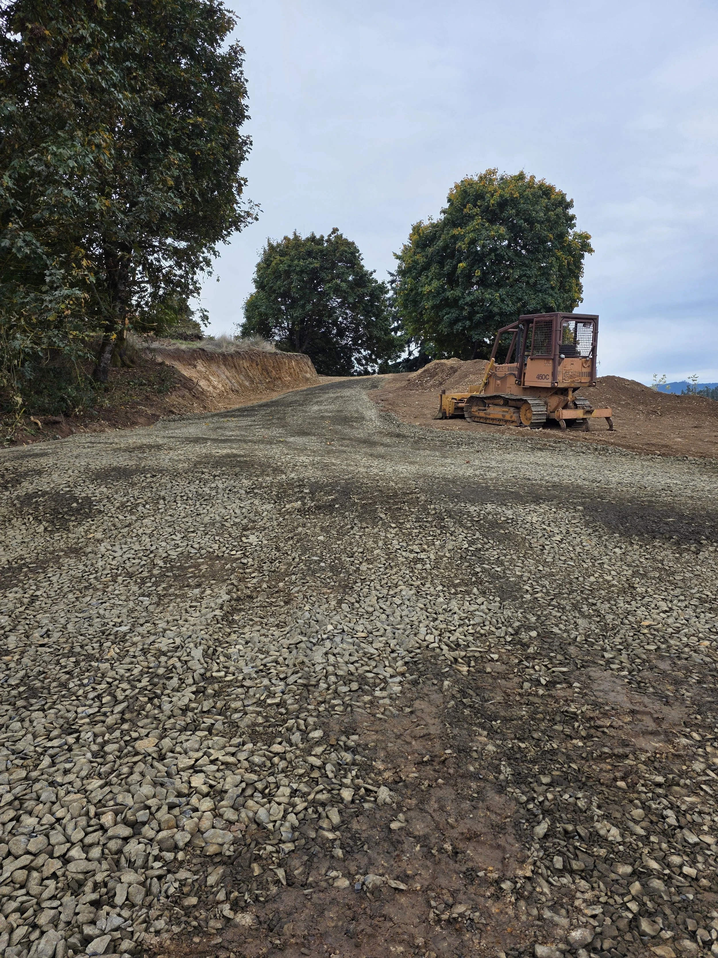 A small construction vehicle parked on a dirt and gravel road surrounded by trees under a cloudy sky.