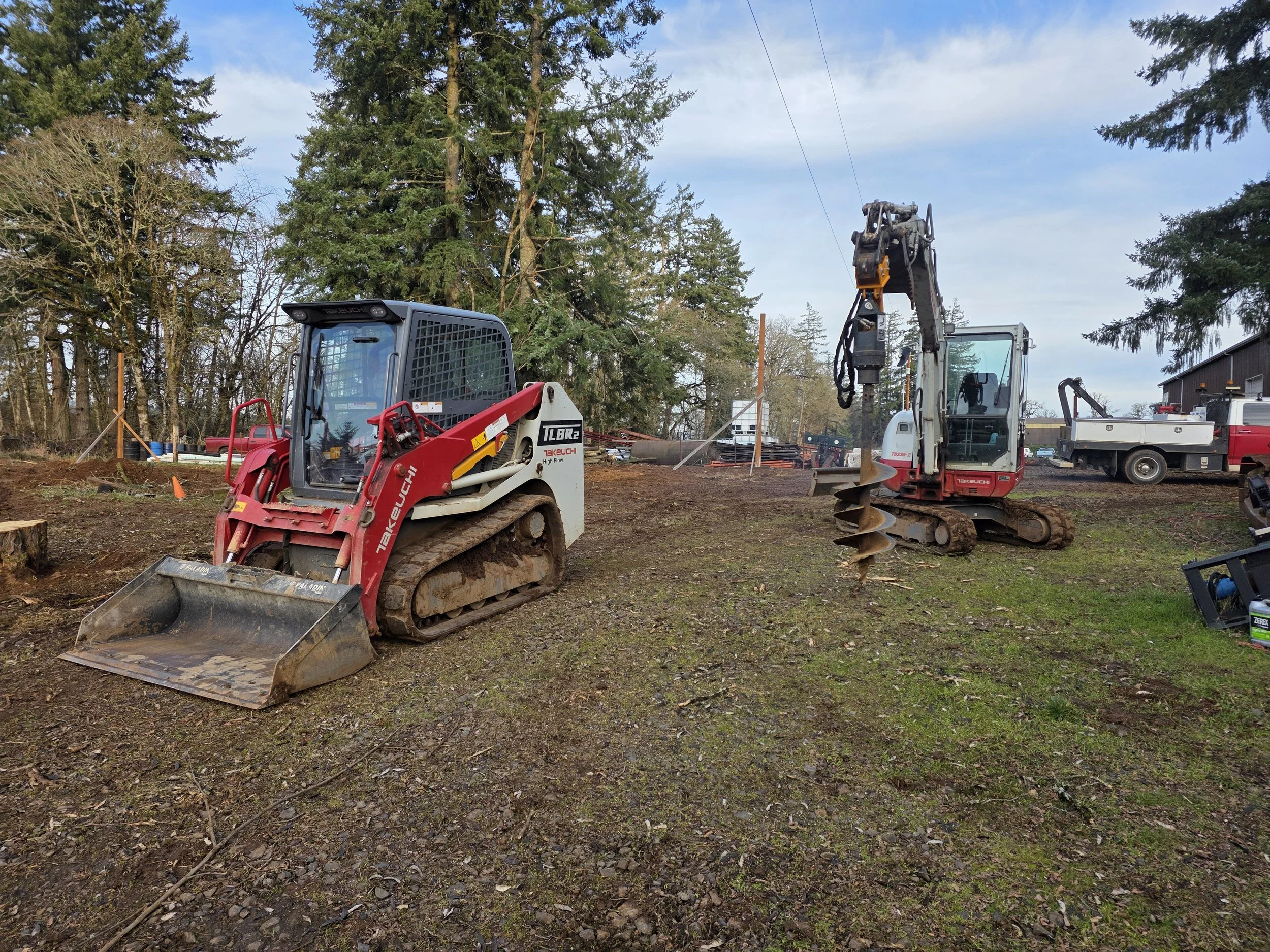 Construction site with a red compact excavator and a white mini excavator, surrounded by trees and construction equipment. Salem Oregon