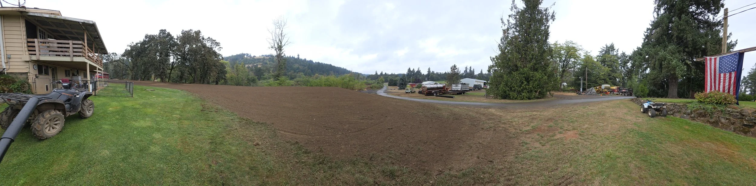 Panoramic view of a rural backyard with a newly tilled garden plot, a house on the left, parked ATVs and other vehicles, a large American flag hanging on a pole, trees, and a cloudy sky.