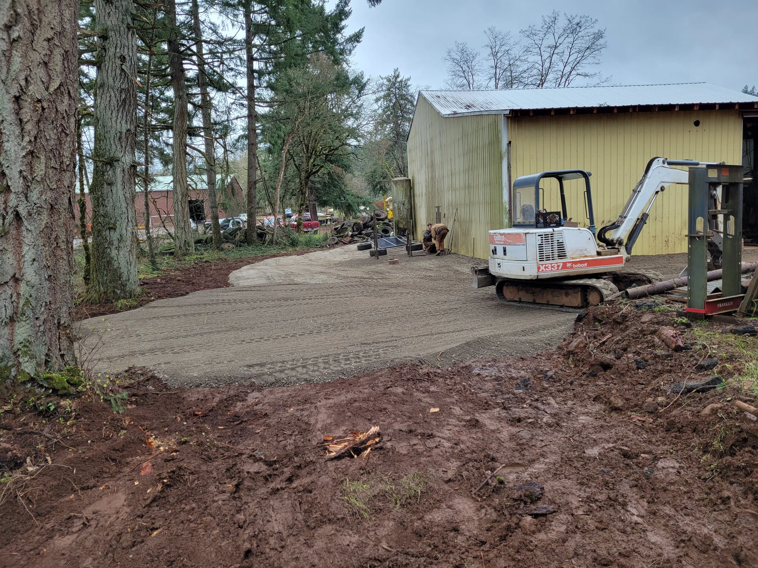 A construction site with a yellow building, trees, a small excavator, and workers. The ground is freshly dug soil, possibly for landscaping or construction. Woodburn Oregon
