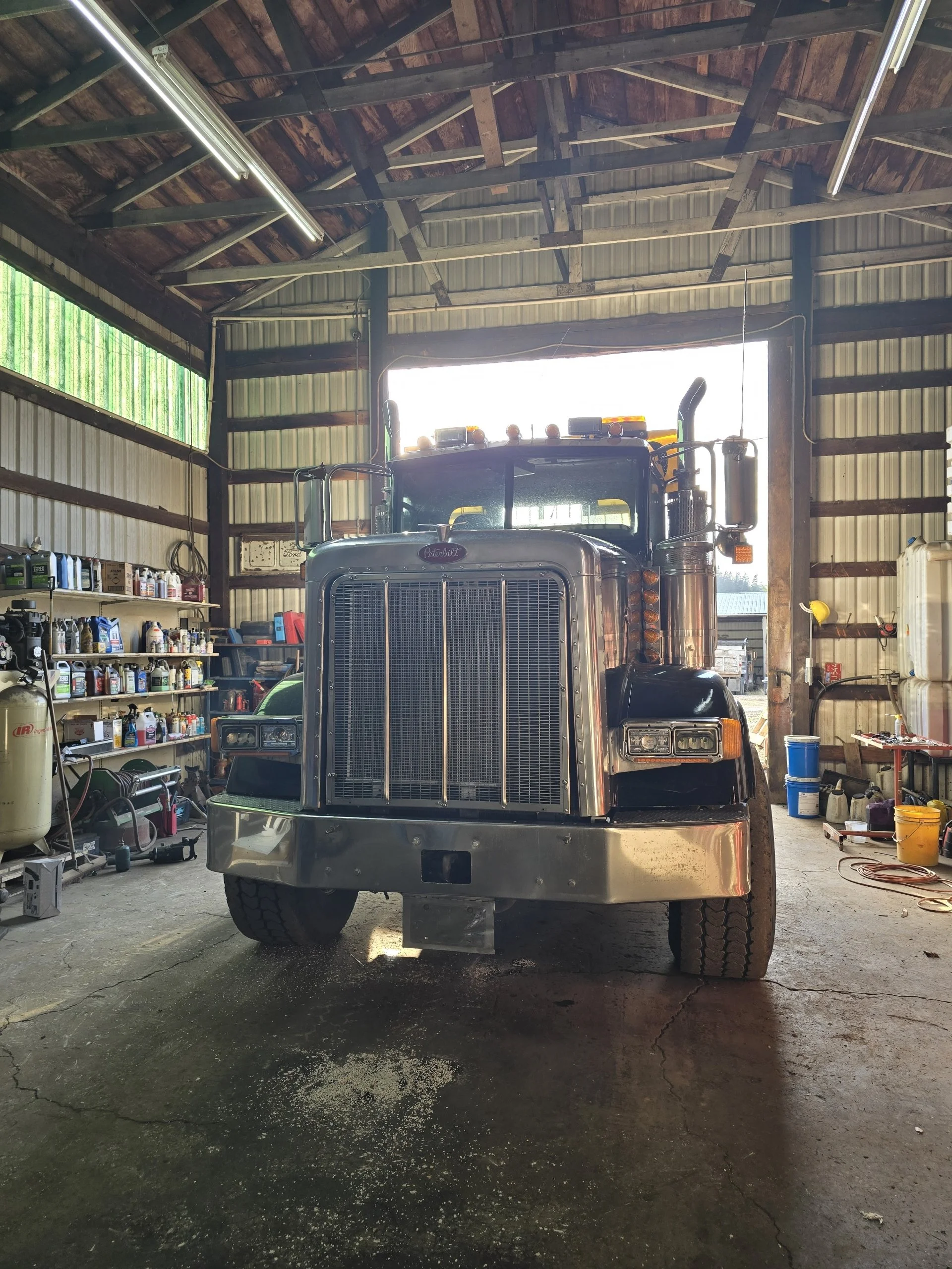 A black semi-truck parked inside a large shed or garage with a metal roof and wooden beams. Shelves on the left side hold various bottles and equipment. The floor shows some water and cracks.