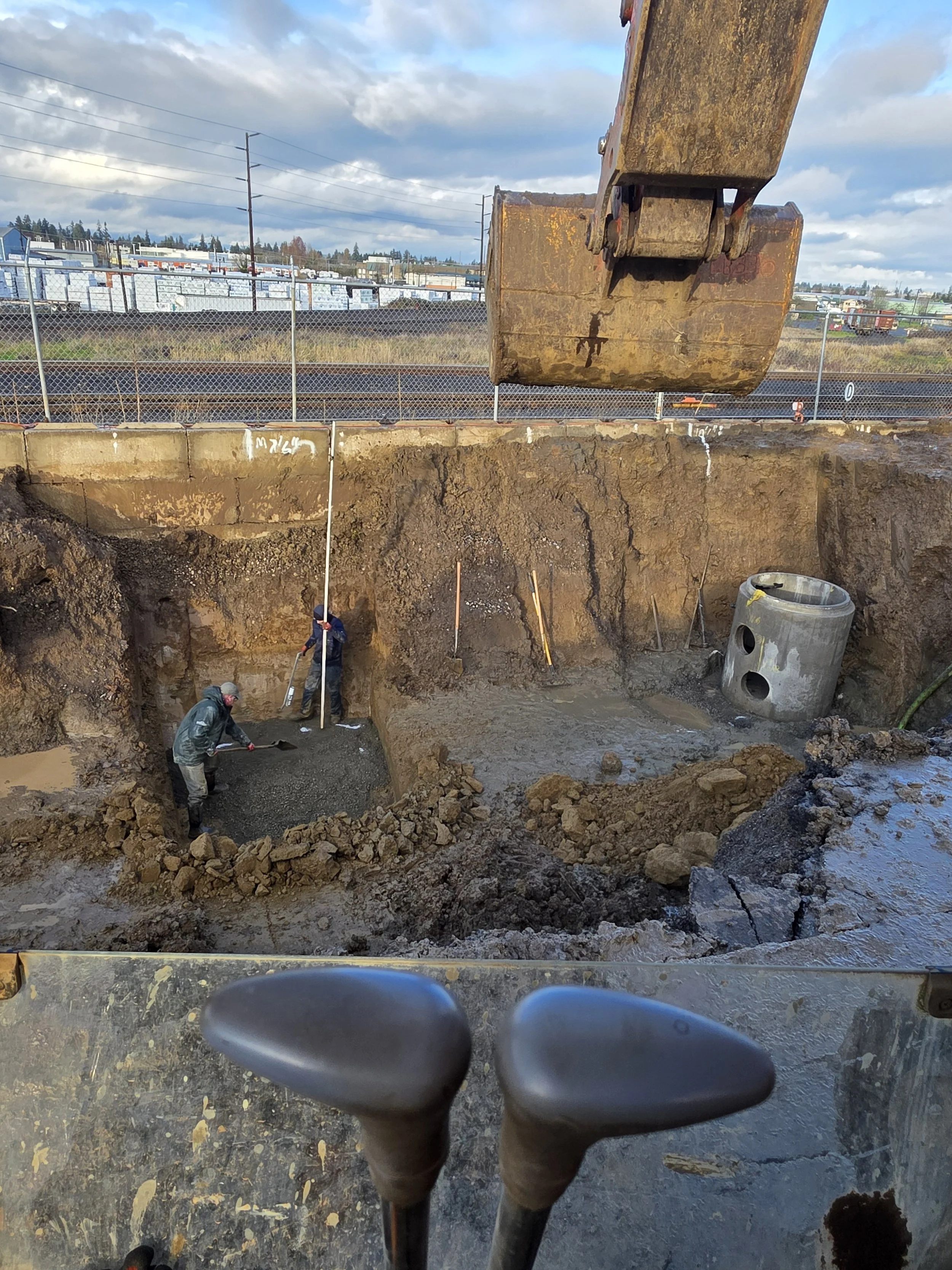 View from bicycle seat looking at construction workers digging a hole with a large excavator arm overhead, with a chain-link fence and industrial buildings in the background under a partly cloudy sky. Albany Oregon