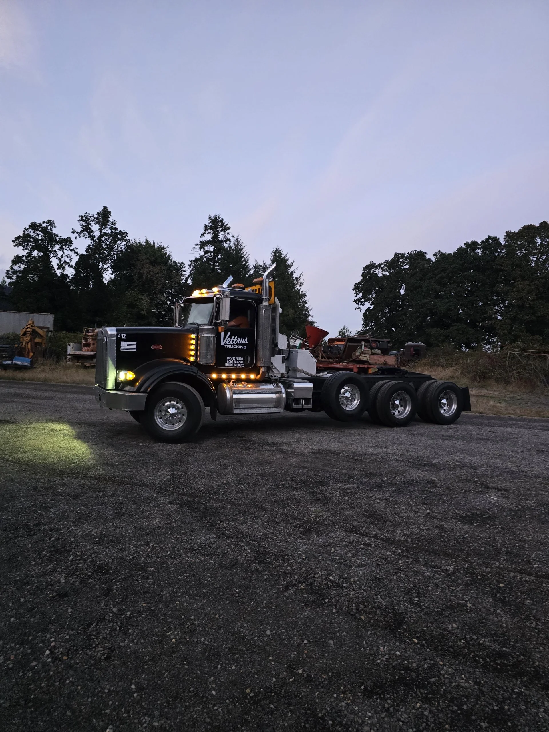 Black semi-truck with yellow lights driving on a gravel road at dusk, with trees in the background.