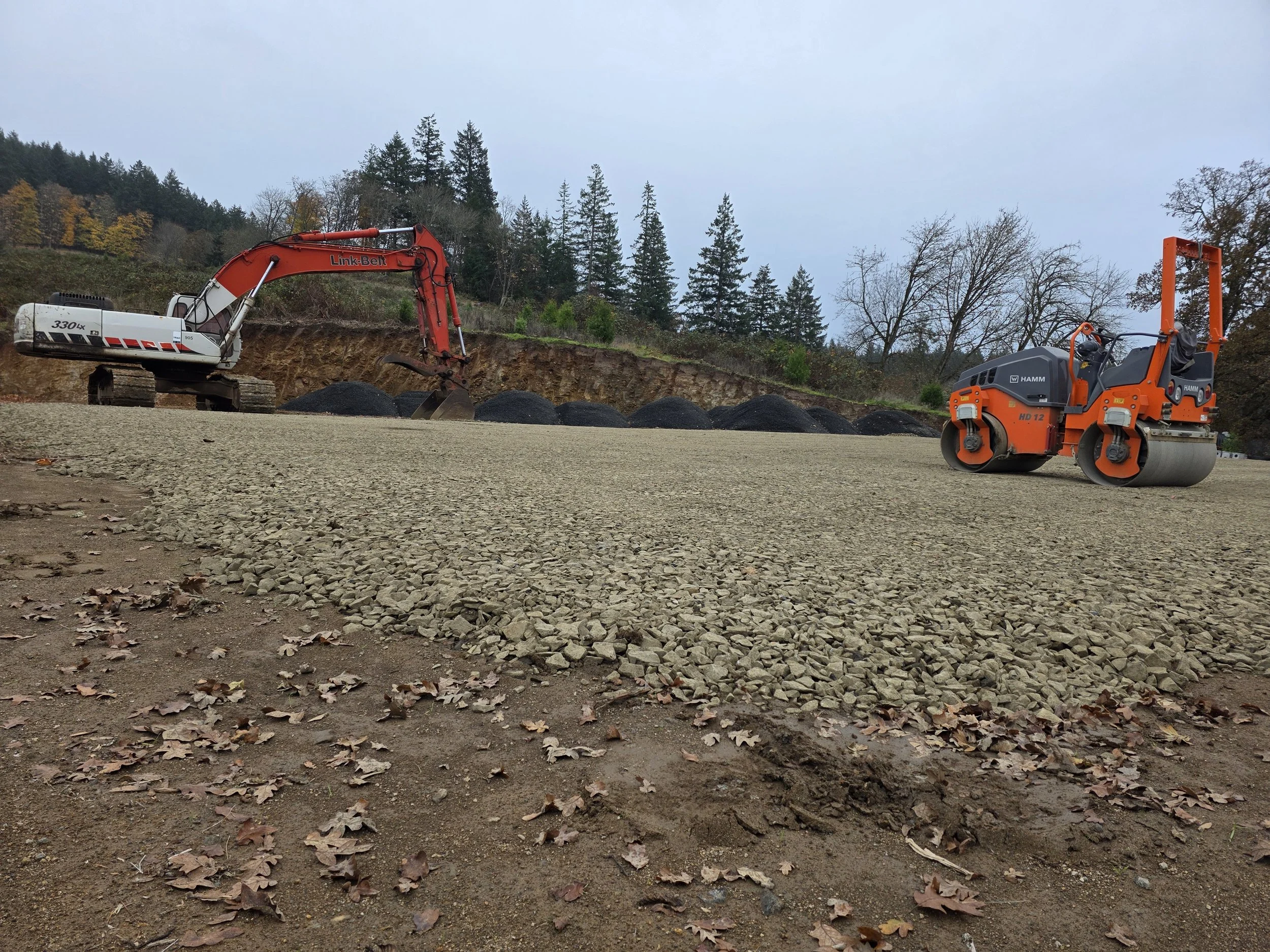 Construction site with an excavator and a road roller on a dirt and gravel surface, with trees and a cloudy sky in the background.