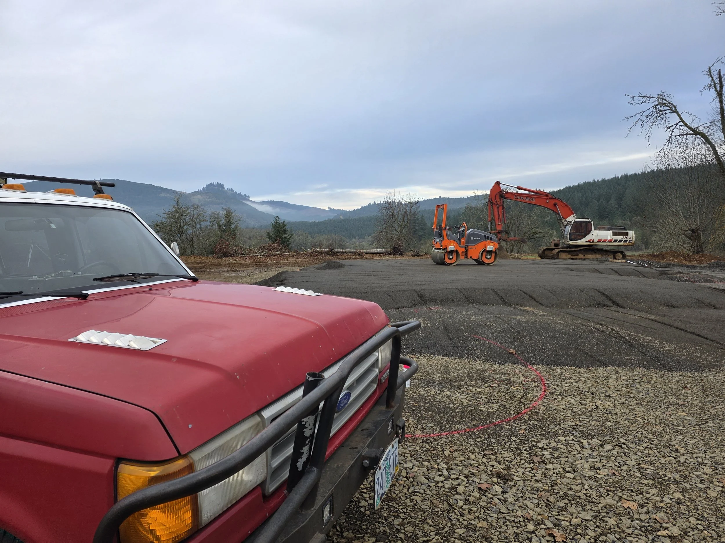 A red Ford vehicle in the foreground. Behind it, there is construction equipment, including an orange compact excavator and a white excavator. The scene is set outdoors with mountains and trees in the background, and the sky is cloudy. doing a house 