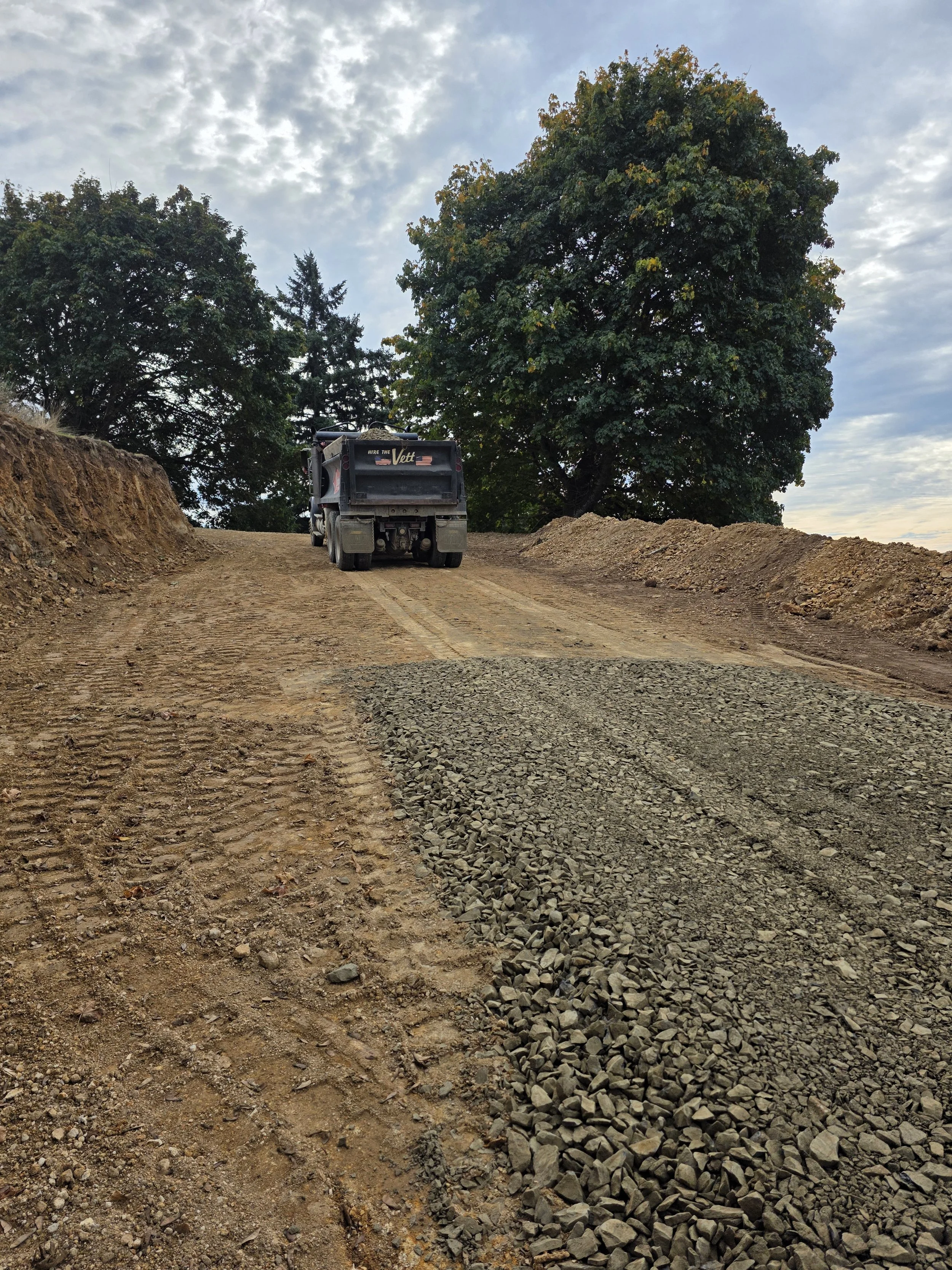 A construction site with a dirt and gravel road, a dump truck, and large trees in the background during daytime with cloudy sky.