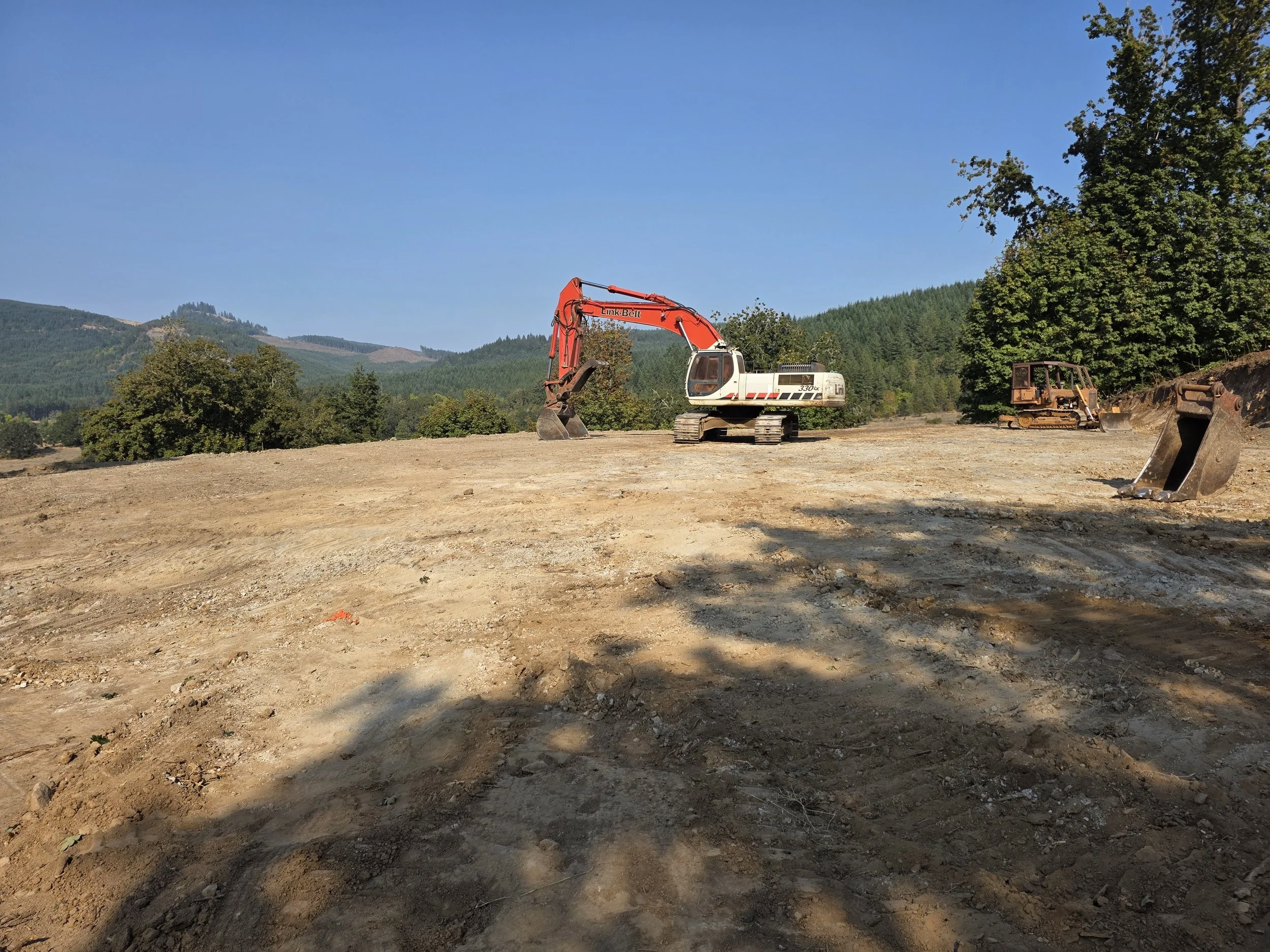 A construction site with a red and white excavator on a dirt field, surrounded by trees and hills under a clear blue sky.