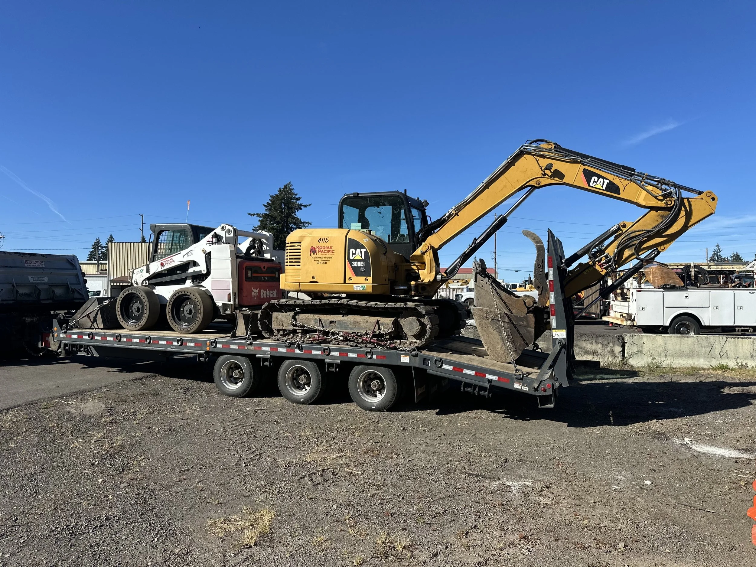 A yellow Caterpillar excavator with black markings on a flatbed trailer, with a small white skid-steer loader loaded next to it, parked in an outdoor lot with other trucks, trees, utility poles, and a clear blue sky in the background.