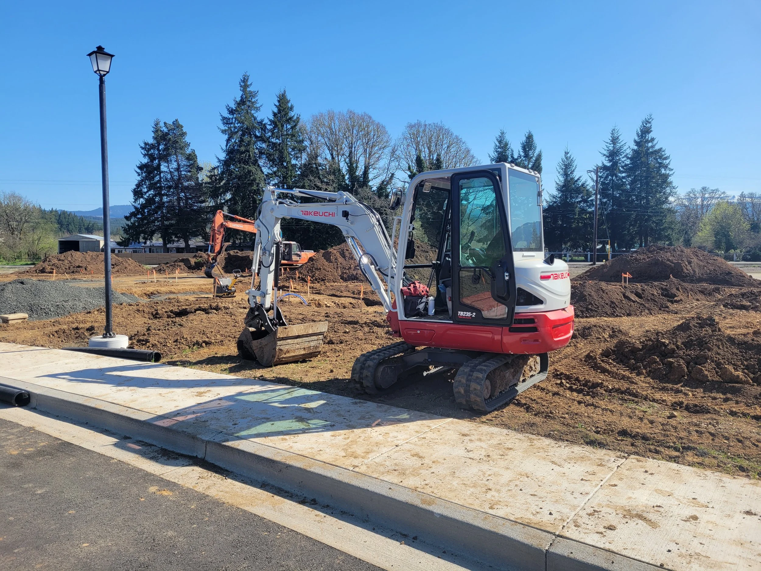 Construction site with a small white and red excavator, large piles of dirt, and construction markings on the ground, against a backdrop of trees and a clear blue sky.