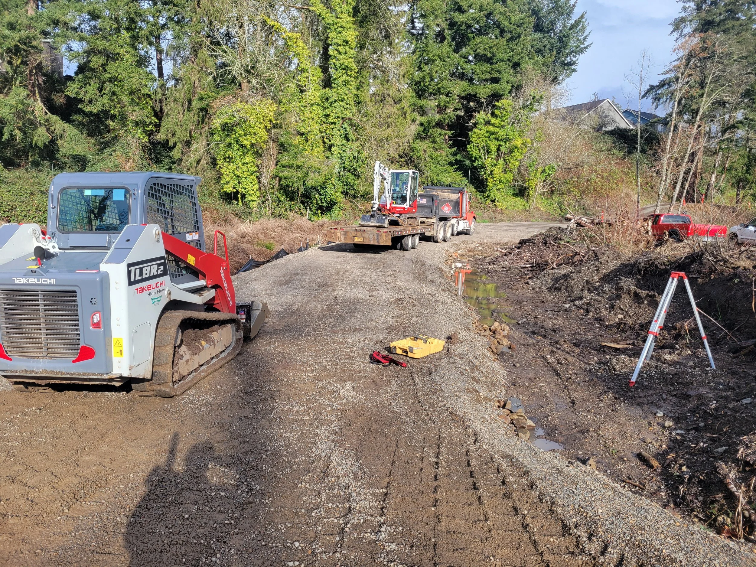 Scene of roadside construction with trees and a house in the background, featuring a small excavator on a flatbed trailer, a skid-steer loader, and various construction tools and equipment.