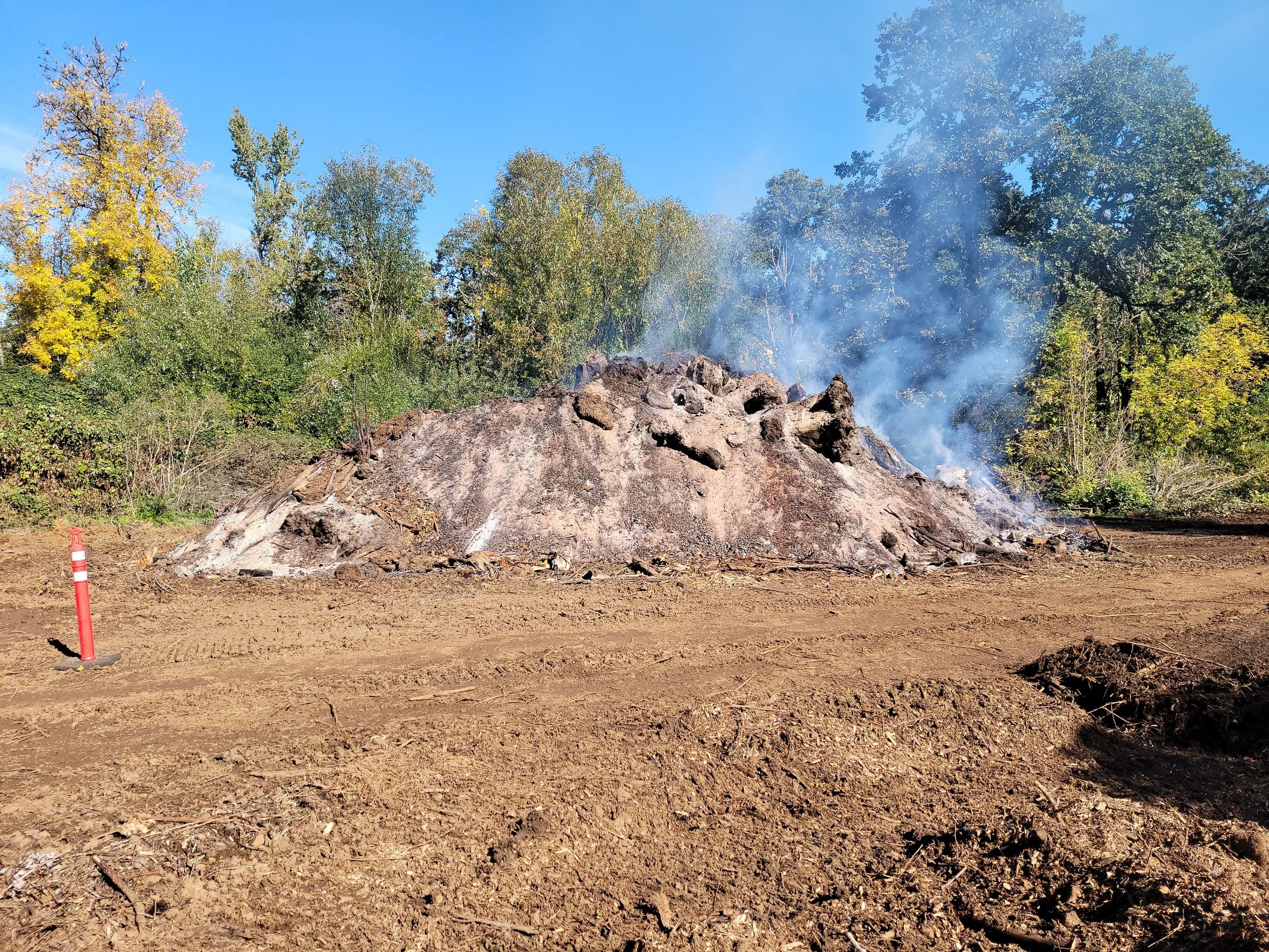 A fire burning in a pile of logs and debris in an outdoor area with trees in the background, blue sky, and smoke rising from the fire. Salem Oregon