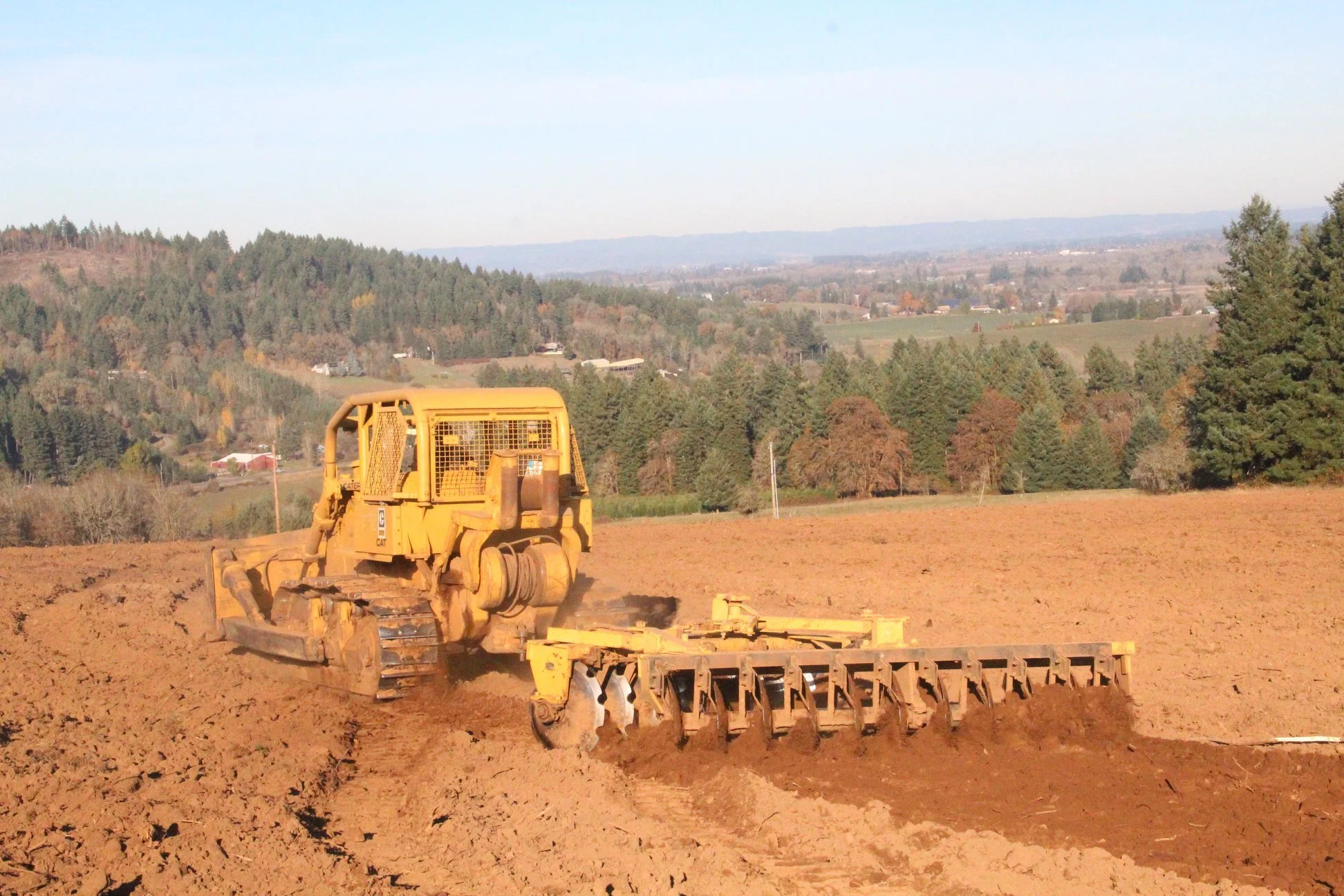 A yellow bulldozer working on a large dirt field with trees and rolling hills in the background. Gaston Oregon