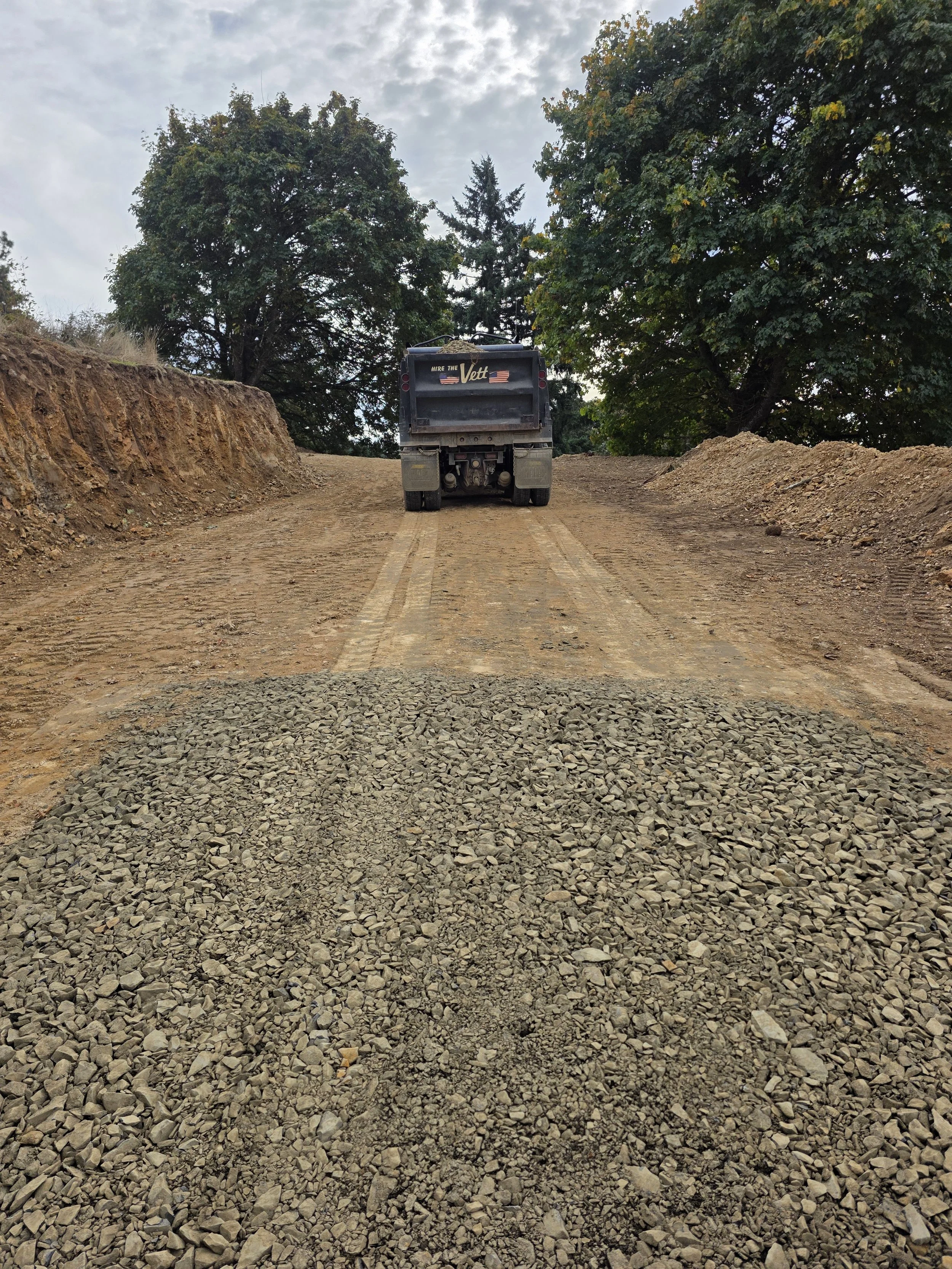 A construction site with a bulldozer on a dirt road, surrounded by trees and cloudy sky.
