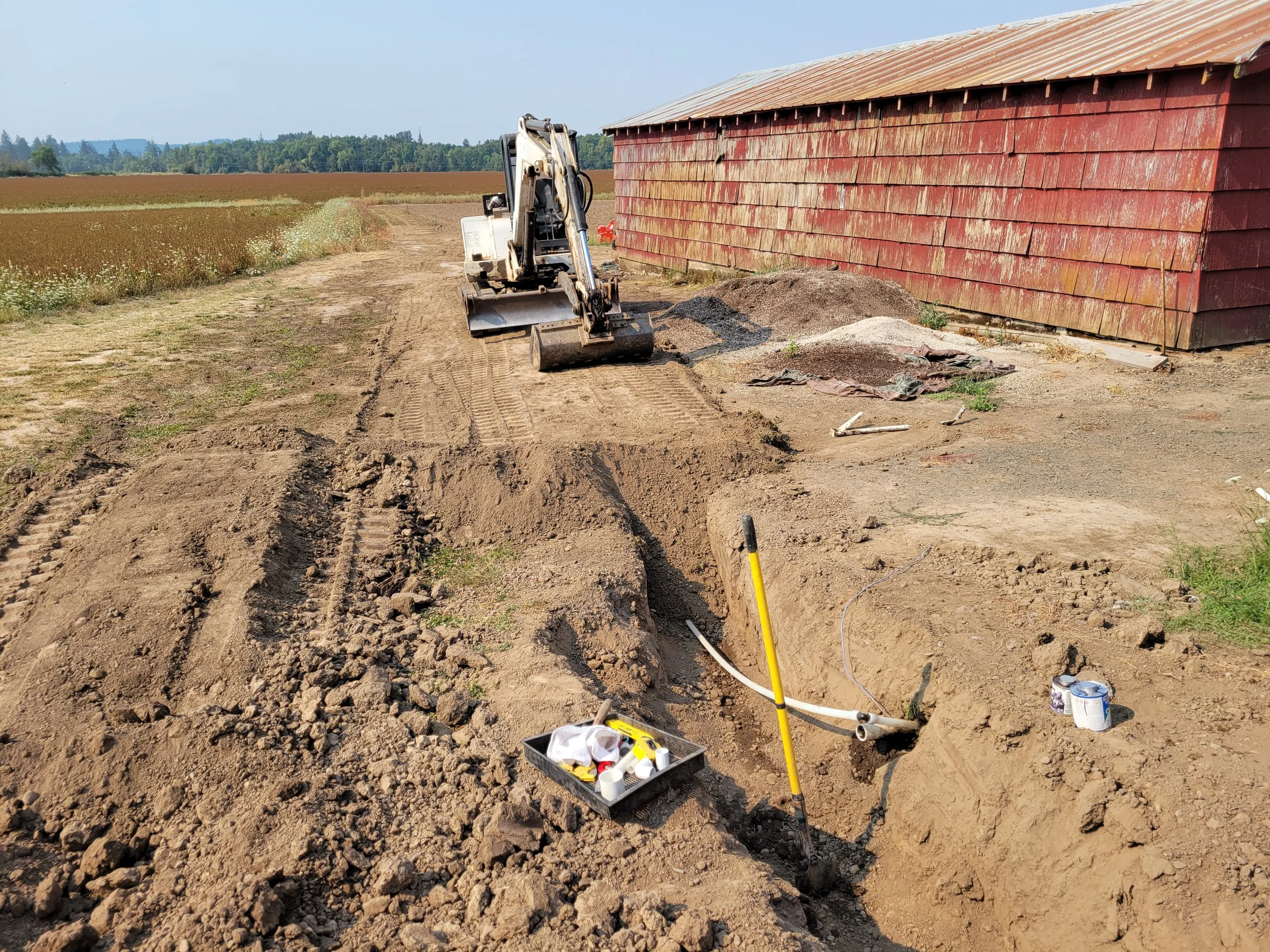 A small excavator parked on a dirt path beside a red wooden barn. In the foreground, there are construction materials, tools, and a yellow measuring pole, with a black plastic container filled with small tubes or bottles. The background shows open fa