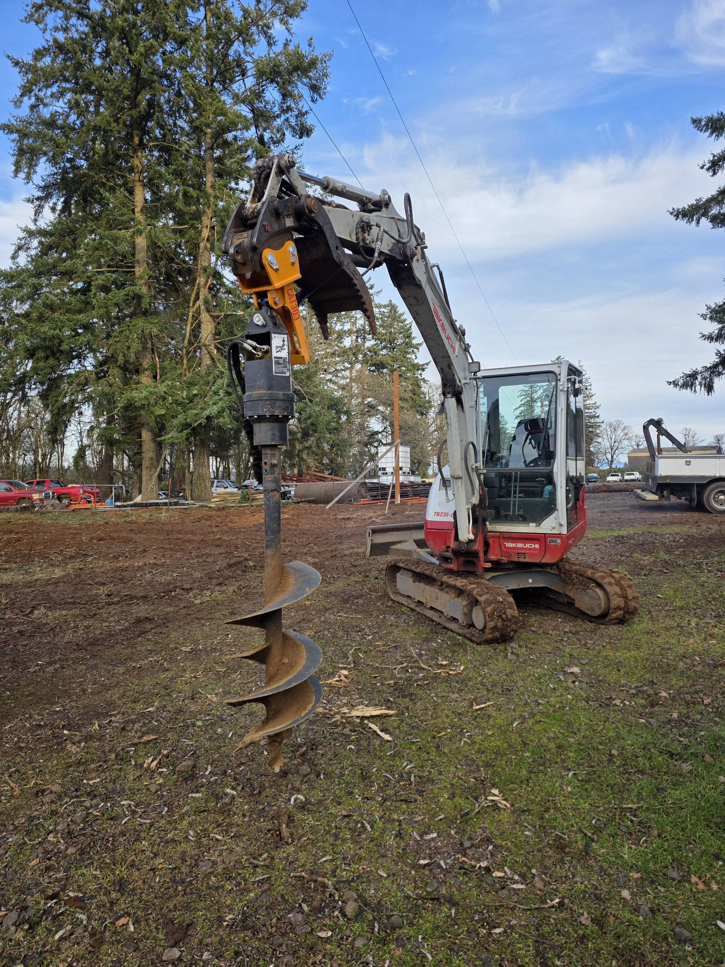 A small excavator with a large auger attachment drilling into the ground at a construction site with trees, a fence, and parked trucks in the background. Aumsville Oregon
