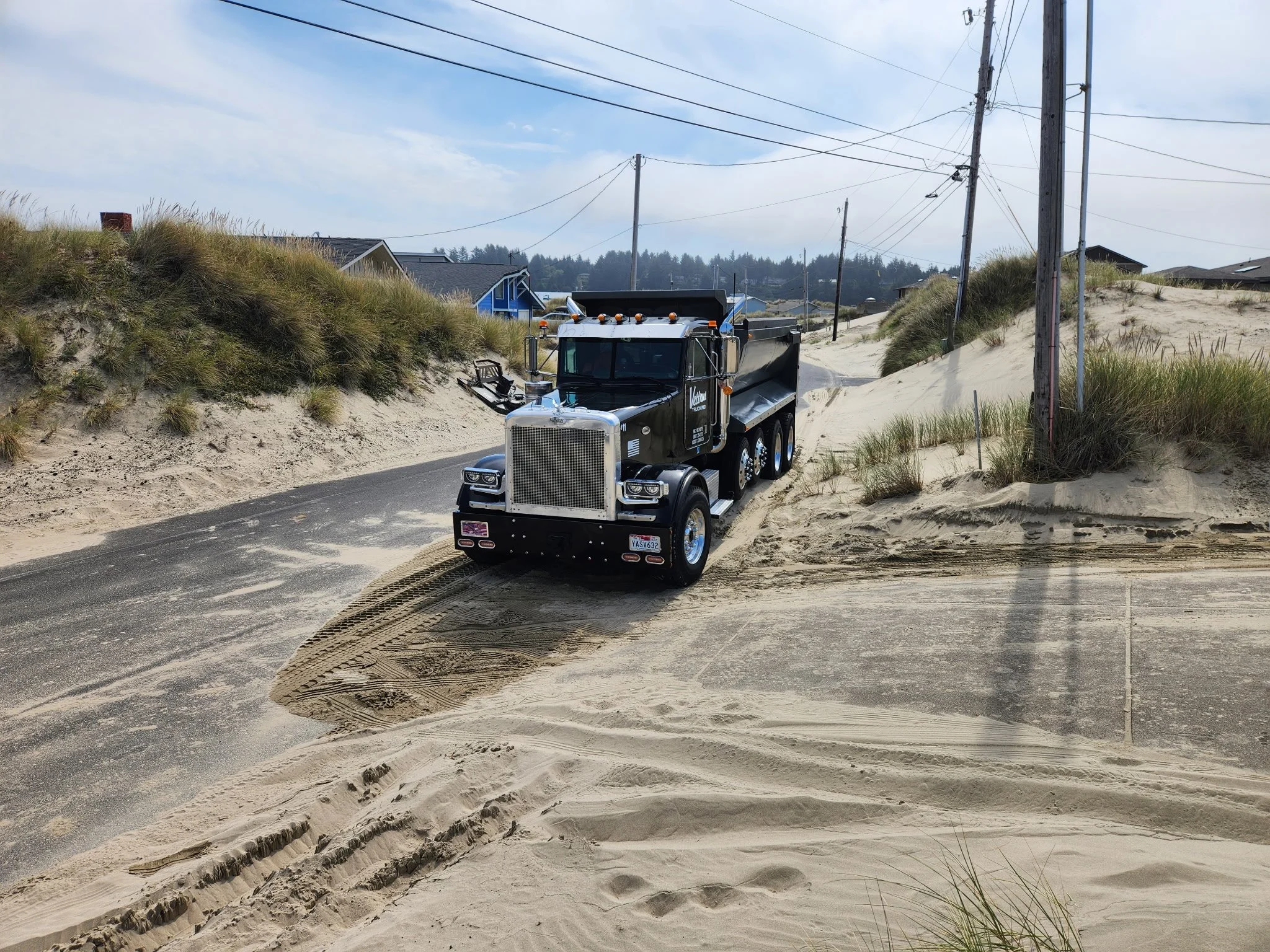 A large black dump truck driving on a road near sandy dunes and grassy terrain with houses and utility poles in the background. The sky is partly cloudy.