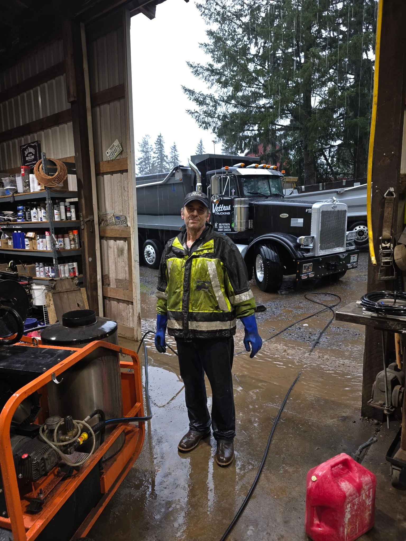 A man wearing waterproof gear and boots standing inside a workshop with a flooded floor. Outside, a large truck is parked, and trees are visible in the background through the open entrance.