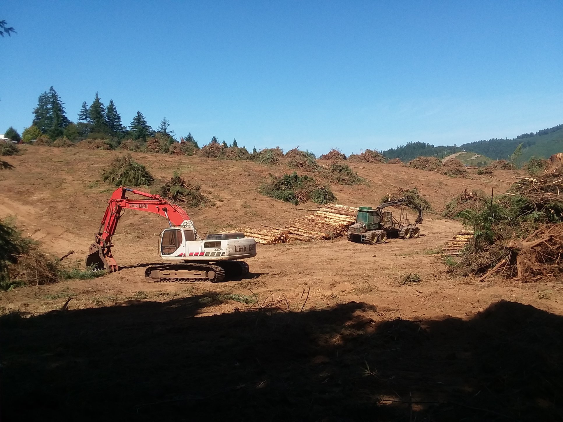 Construction site with heavy machinery, including an excavator and a logging tractor, clearing land under a clear blue sky with trees and hills in the background.