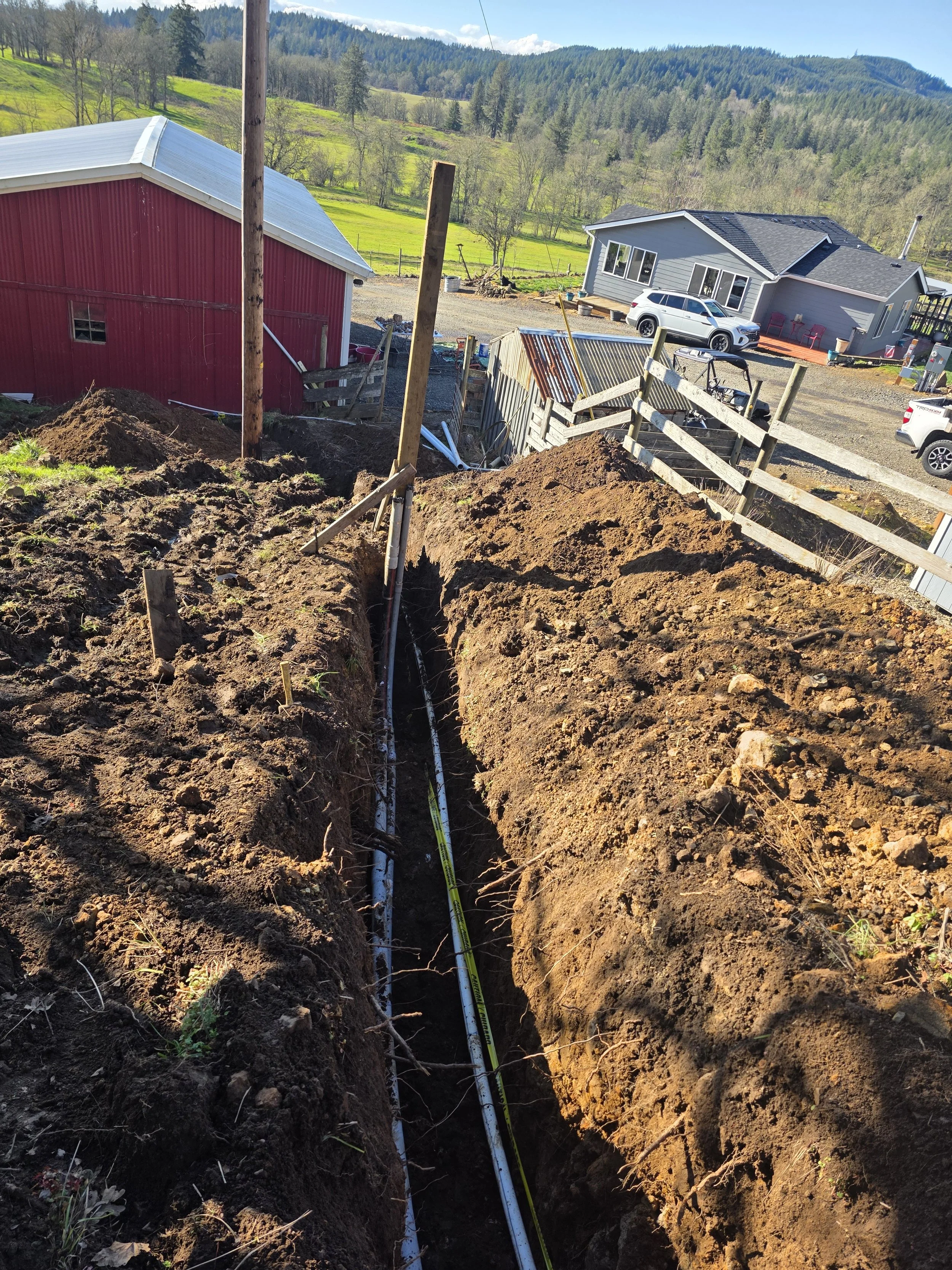 A construction site in a rural area shows a long trench dug in the ground with white pipes laid inside. The scene includes dirt mounds on either side of the trench and wooden posts supporting a partially collapsed fence. In the background, there are 