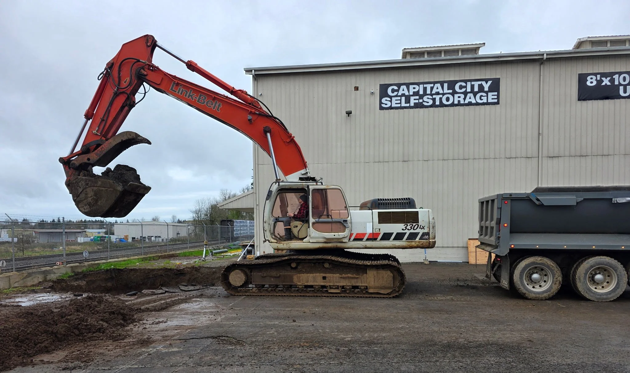 A construction excavator with an orange arm and tracks is digging a hole in the ground at a construction site. The excavator is positioned near a large building with signs advertising self-storage units, and a truck is parked nearby. The scene appear