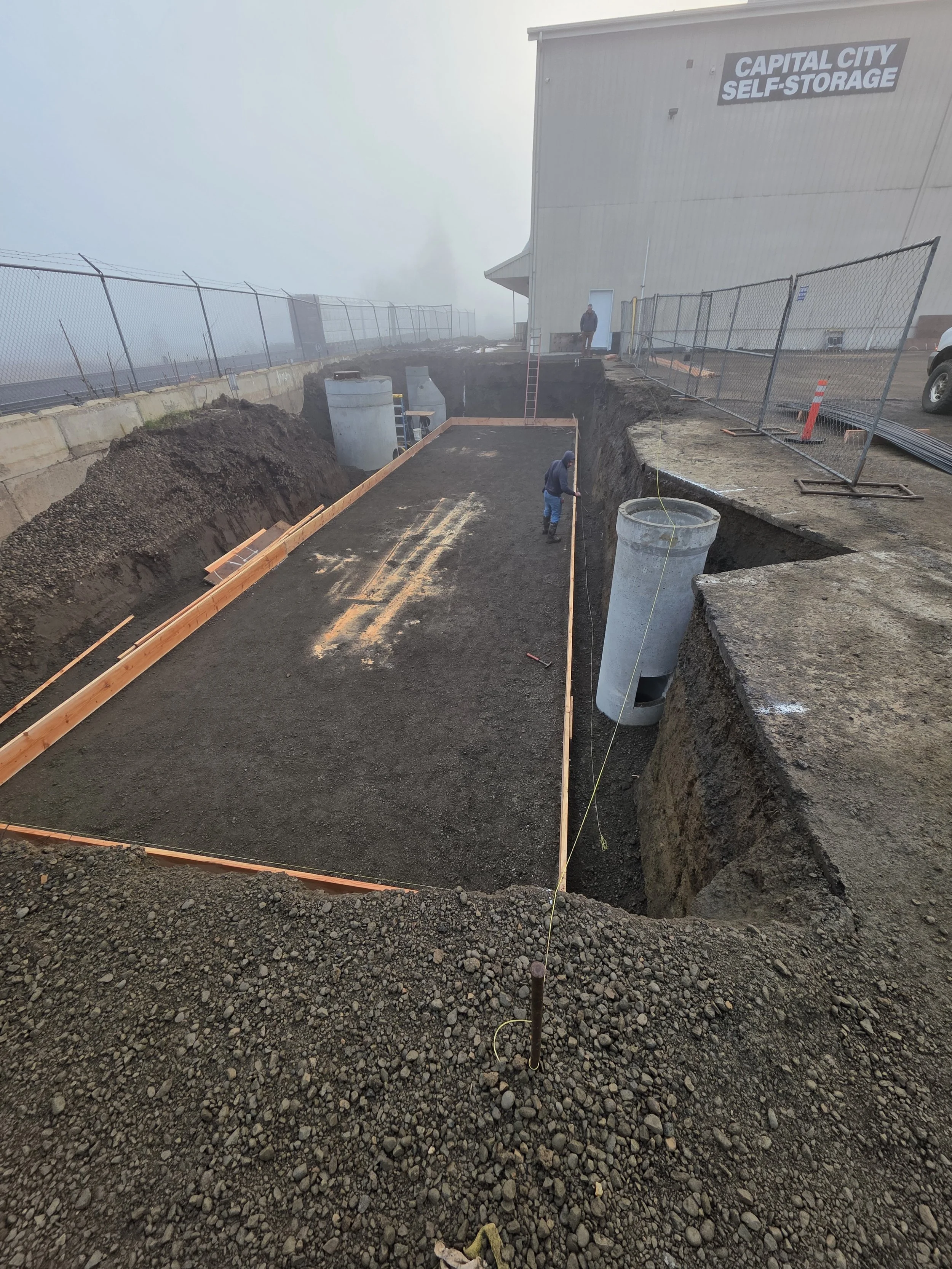 Construction site with a deep trench, wooden forms, and concrete pipes, next to a building with a sign that reads 'CAPITAL CITY SELF-STORAGE'; workers are seen measuring and working on the project.