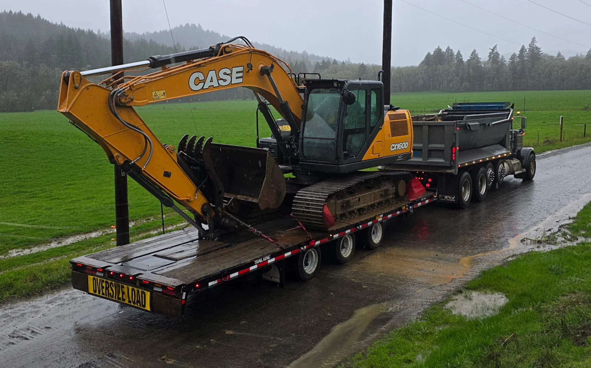 A large yellow CASE excavator loaded onto a flatbed tow truck on a rainy day, with a green field and forested hills in the background. Salem Oregon