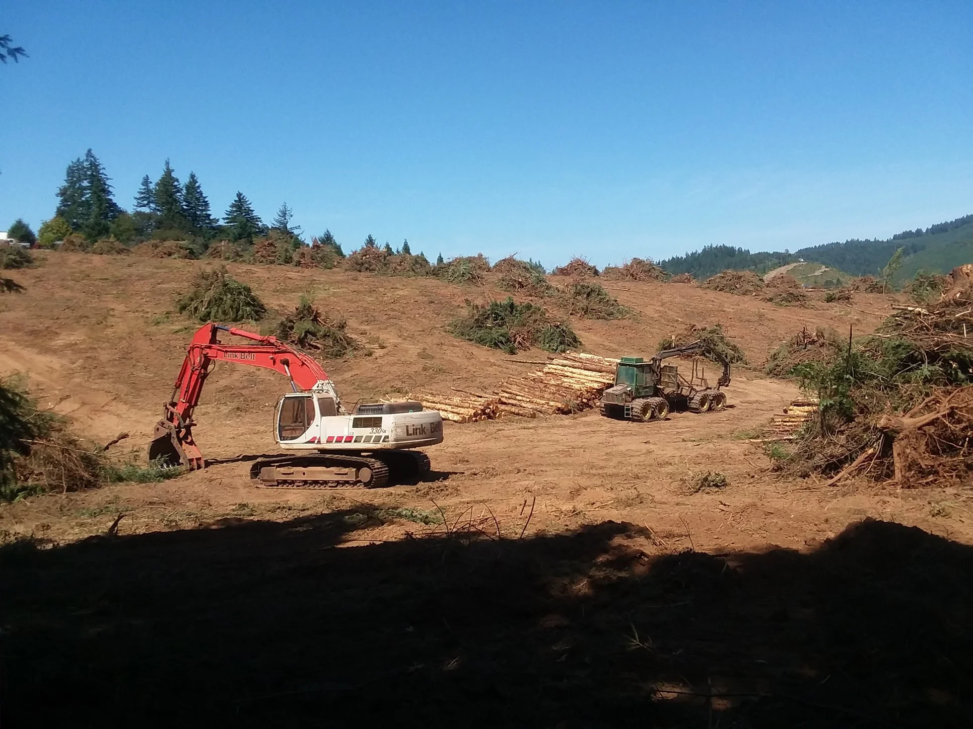 Construction site with excavator and logging equipment on cleared land, hills and trees in the background, partly sunny sky. Gaston Oregon