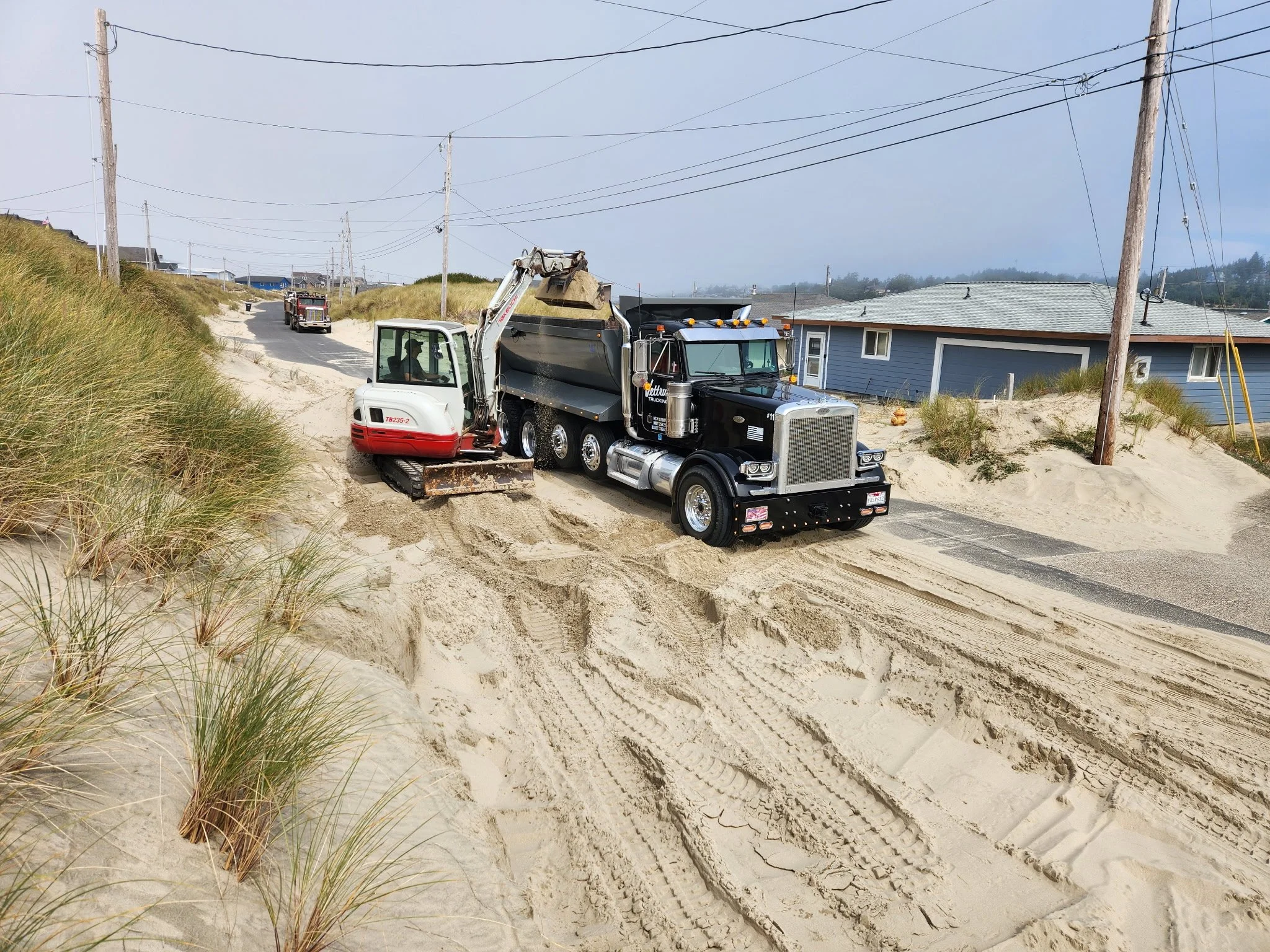 A black semi-truck with a dirt excavator attachment parked in a sandy area near beach dunes and a house, with electrical poles and wires overhead. Lincoln City Oregon