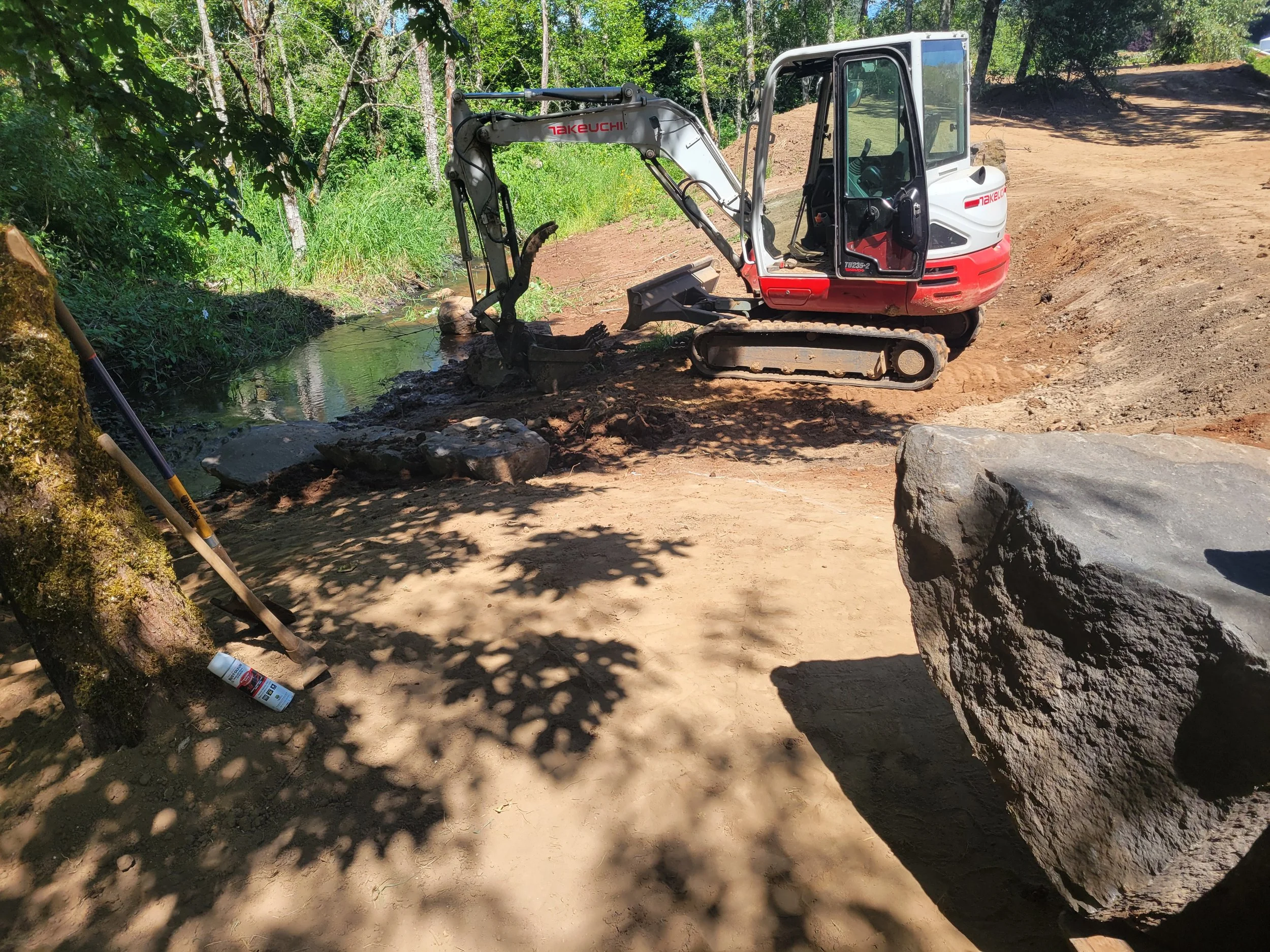 A small white and red mini excavator operating on a dirt construction site near a small stream surrounded by trees and greenery.