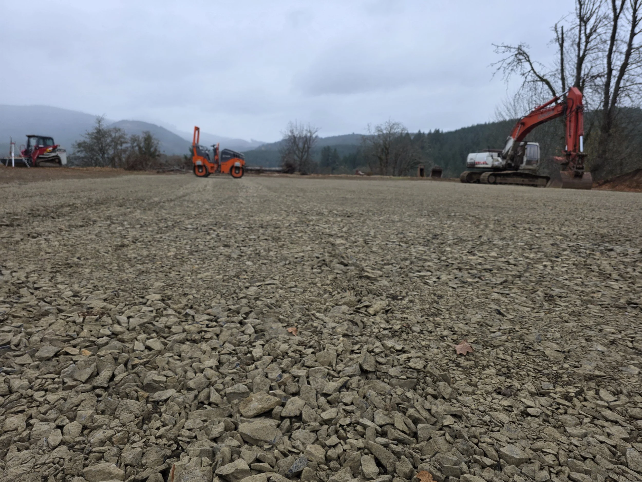 Construction site with gravel ground, two small excavators, and a small bulldozer, with trees and mountains in the background under cloudy sky.