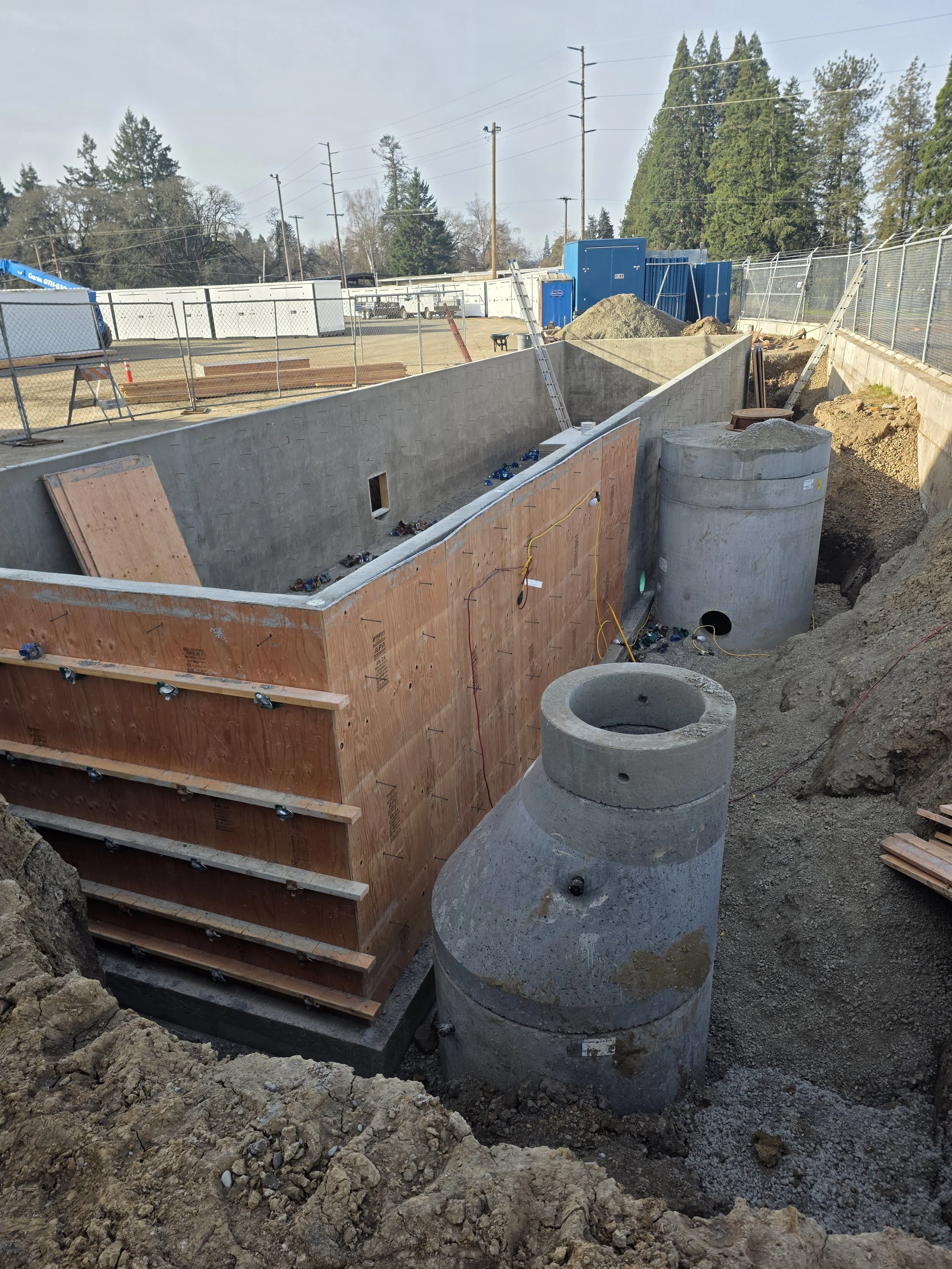 Construction site with large concrete pipes, wooden formwork, dirt, and fencing, indicating ongoing infrastructure work. Dallas Oregon
