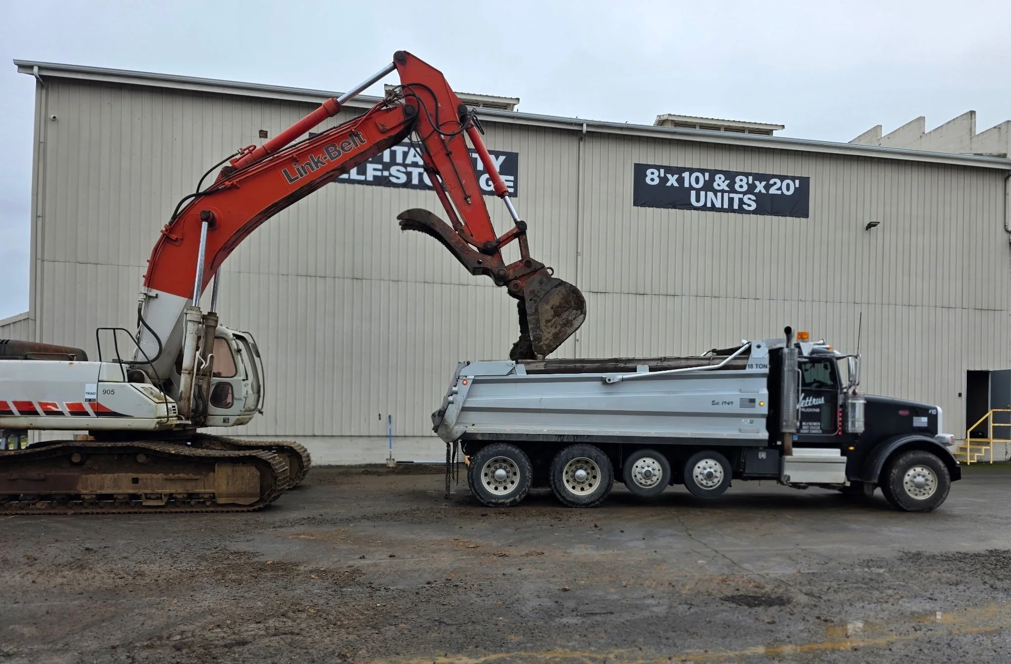 An orange hydraulic excavator with a bucket attachment lifting dirt into a white dump truck outside a large industrial building.