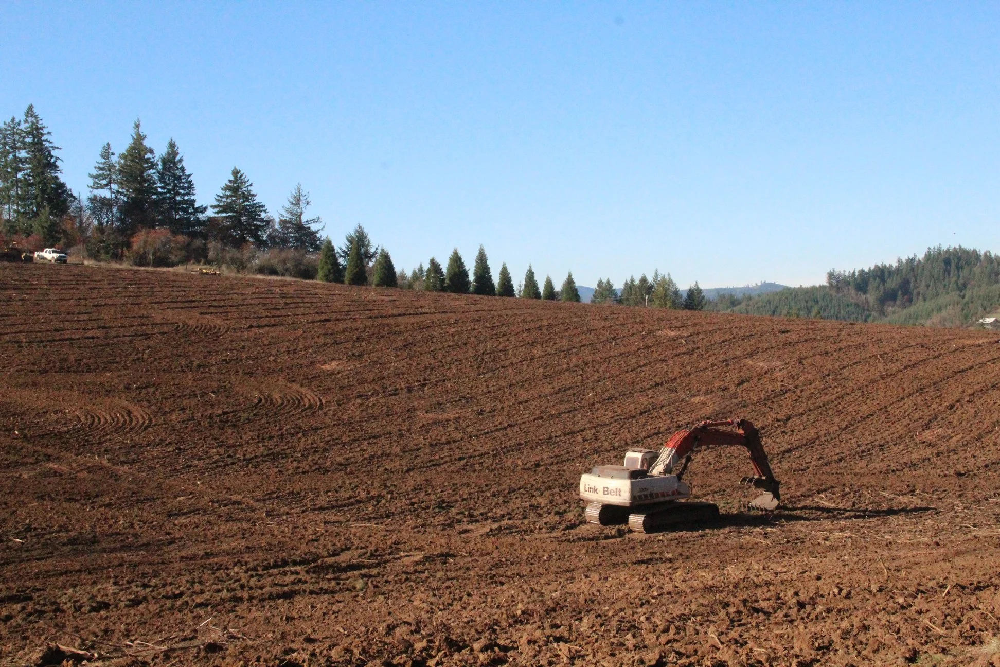 A construction site with freshly tilled earth and a small excavator in the foreground, with rolling hills and a treeline under a clear blue sky in the background.
