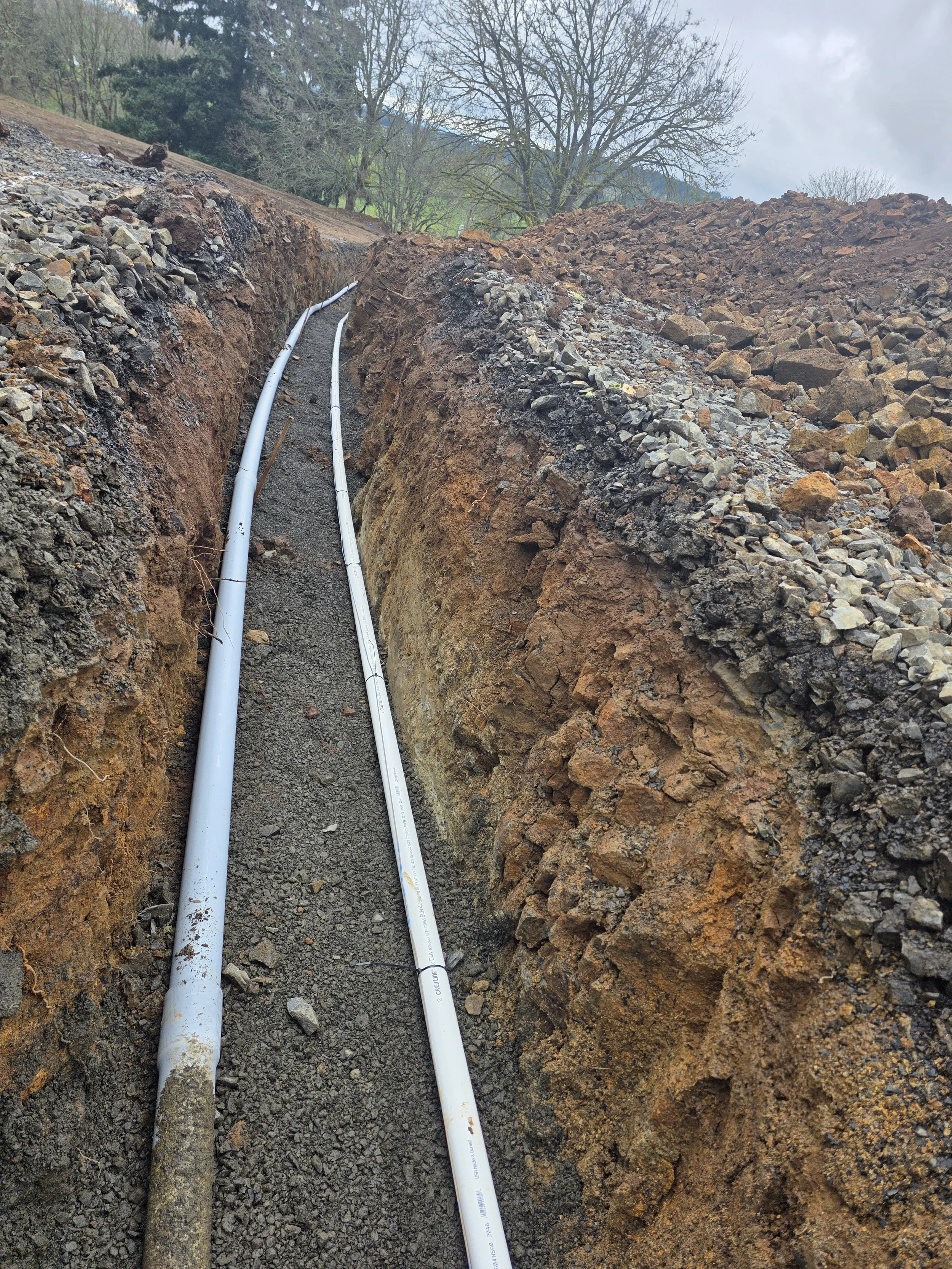 Two white pipes laid in a trench dug into the earth, with one pipe damaged, surrounded by rocks and dirt, with trees and cloudy sky in the background. Digging utilities Corvallis Oregon.